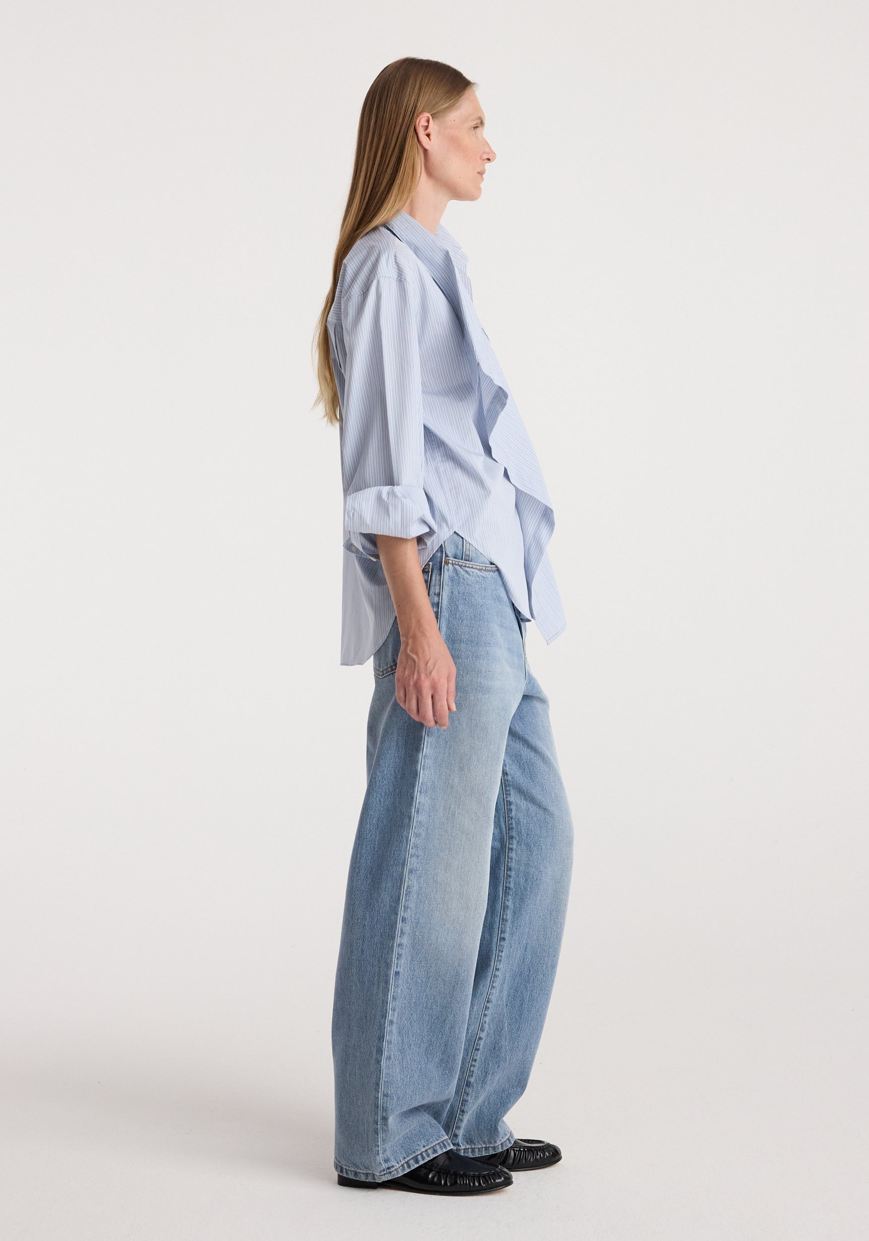 Woman wearing blue striped Draped collar shirt | blue stripe paired with light blue wide-leg jeans and black shoes, standing in profile against a plain background.