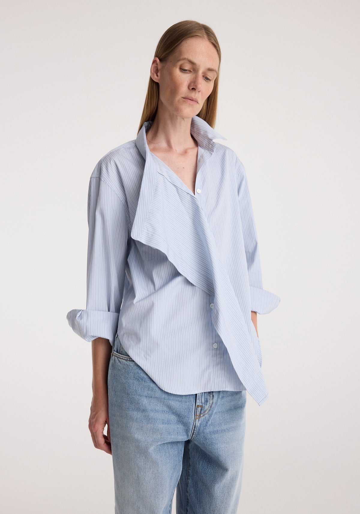 Woman wearing a blue and white striped draped collar shirt | blue stripe paired with light blue jeans, standing against a plain background with hands in pockets.