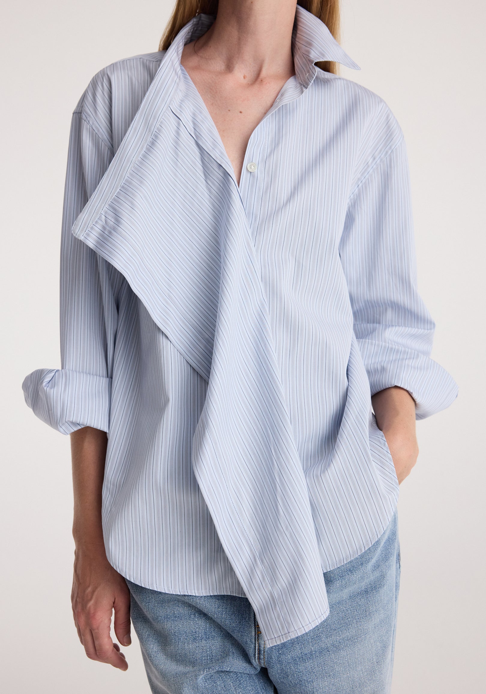 Woman wearing a draped collar shirt | blue stripe with rolled-up sleeves and light blue jeans, showcasing the unique draped design and fine vertical stripes.