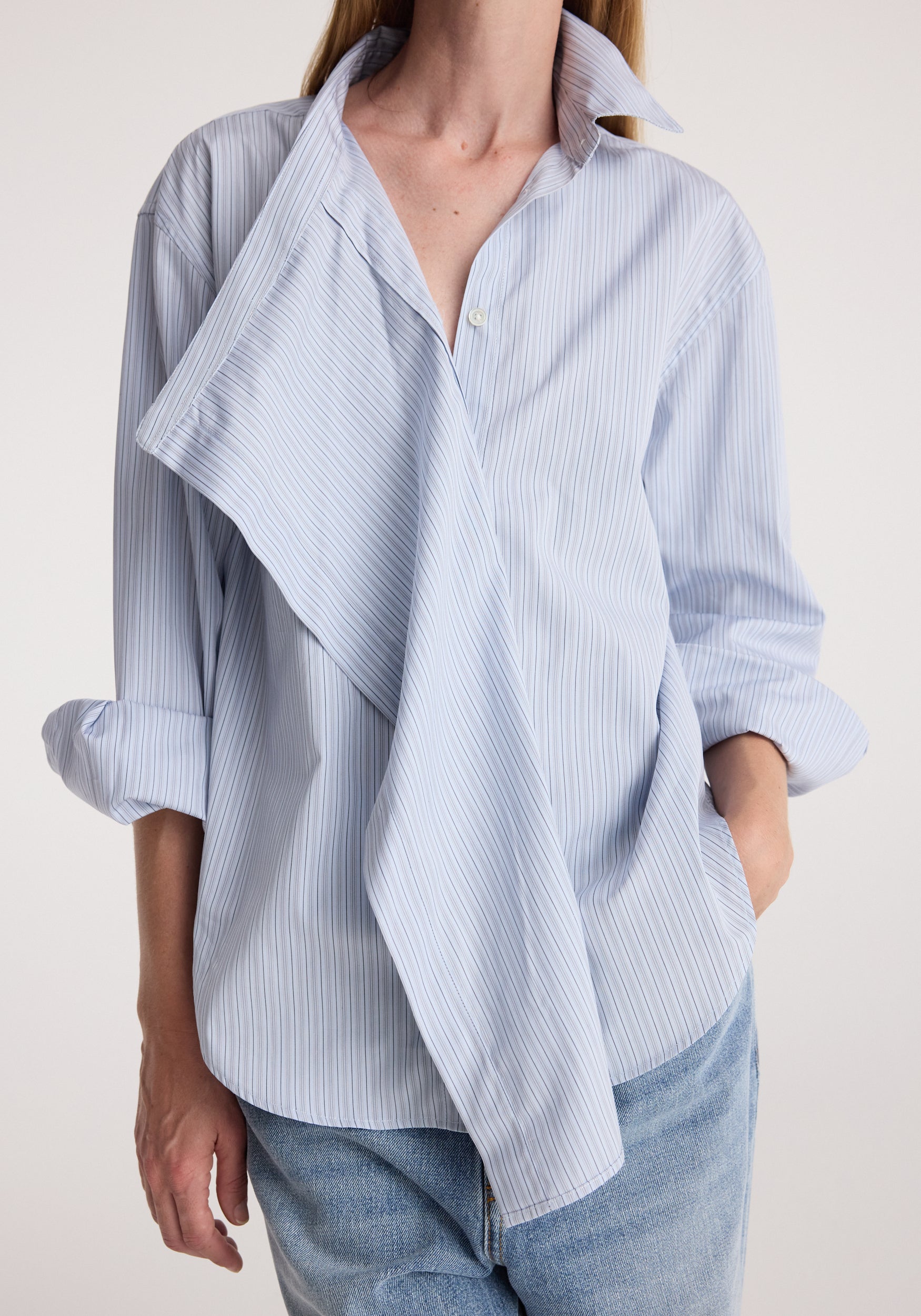 Woman wearing a draped collar shirt | blue stripe with rolled-up sleeves and light blue jeans, showcasing the unique draped design and fine vertical stripes.