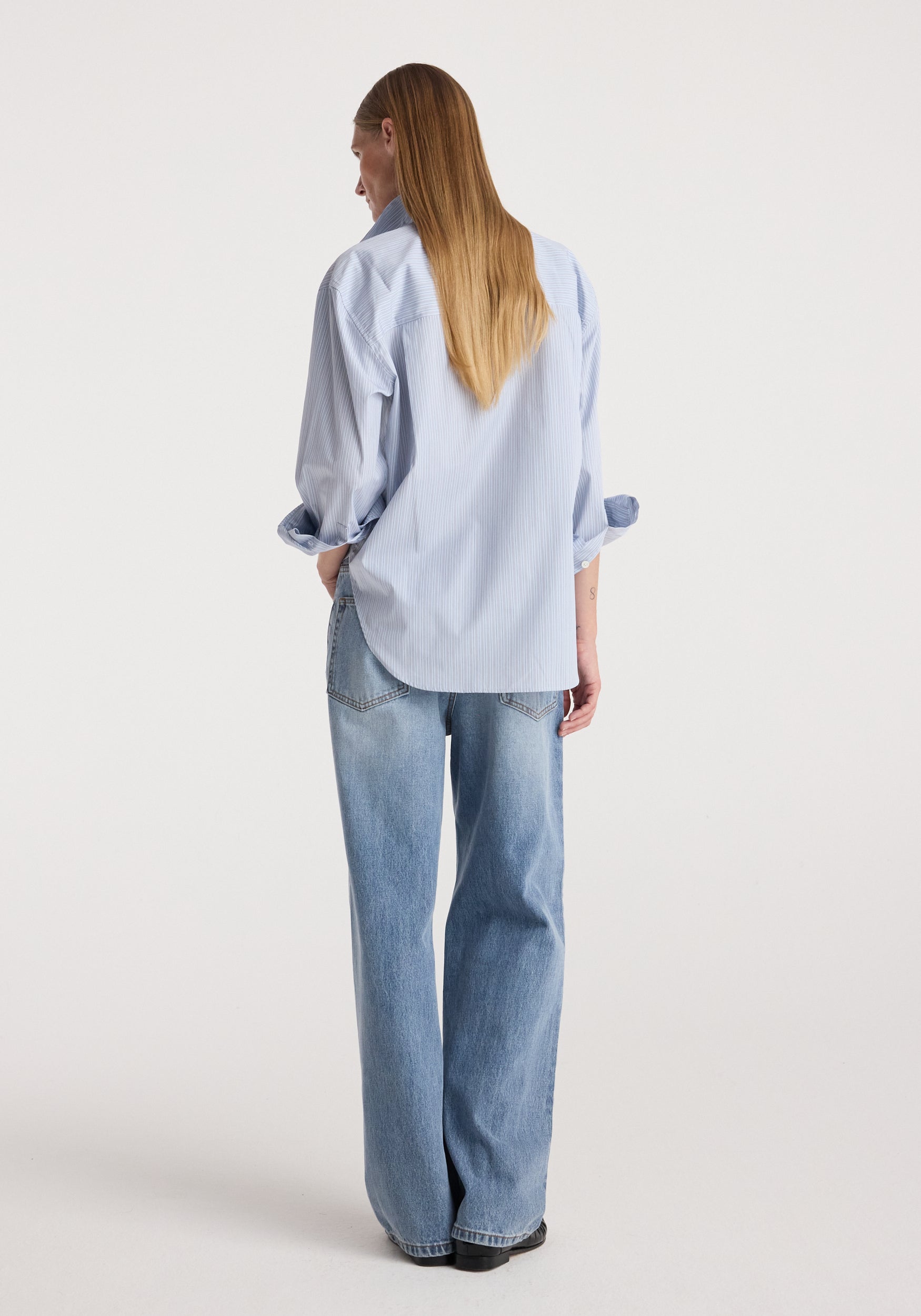 Woman wearing light blue striped Draped collar shirt | blue stripe with rolled-up sleeves and relaxed fit, paired with loose blue jeans, facing away on white background.