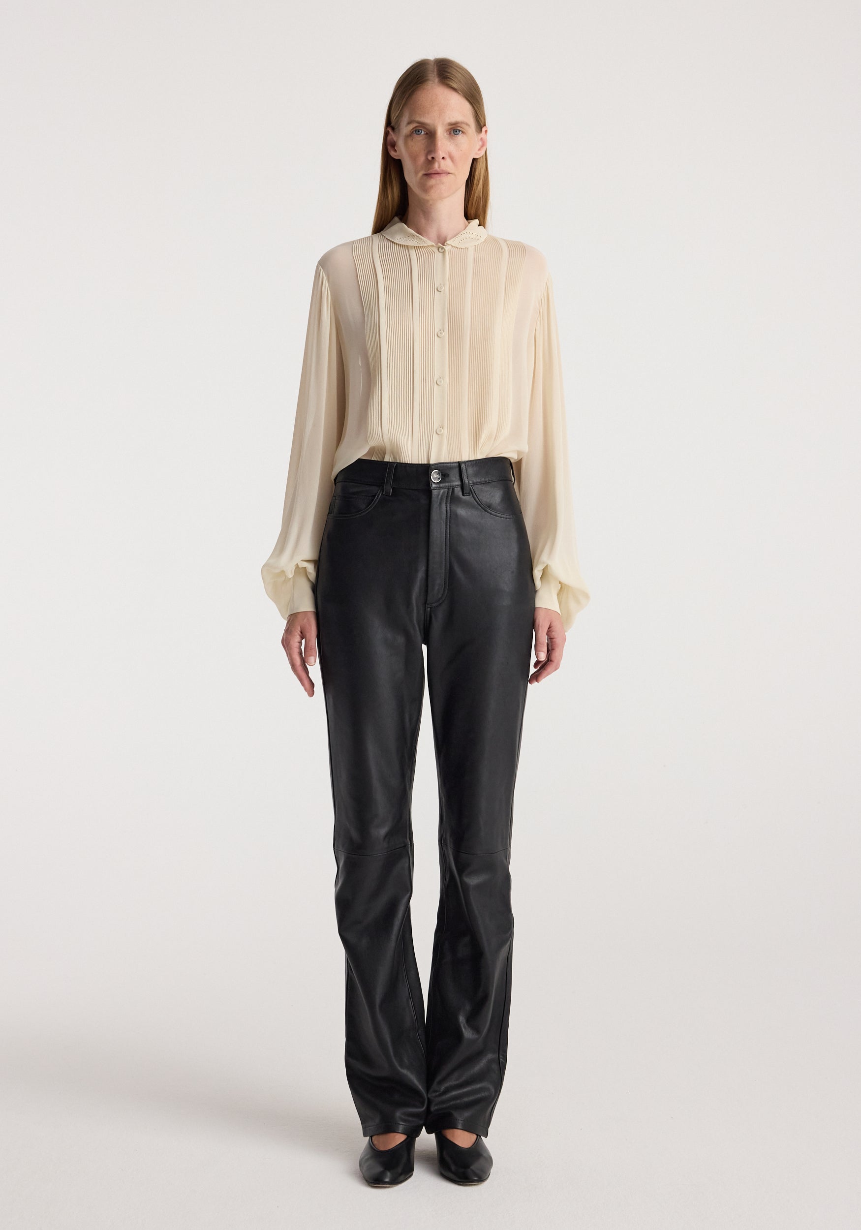 Woman wearing a cream-colored Romantic top | cream with long sleeves and button-up front, paired with black high-waisted pants and black shoes against a plain background.