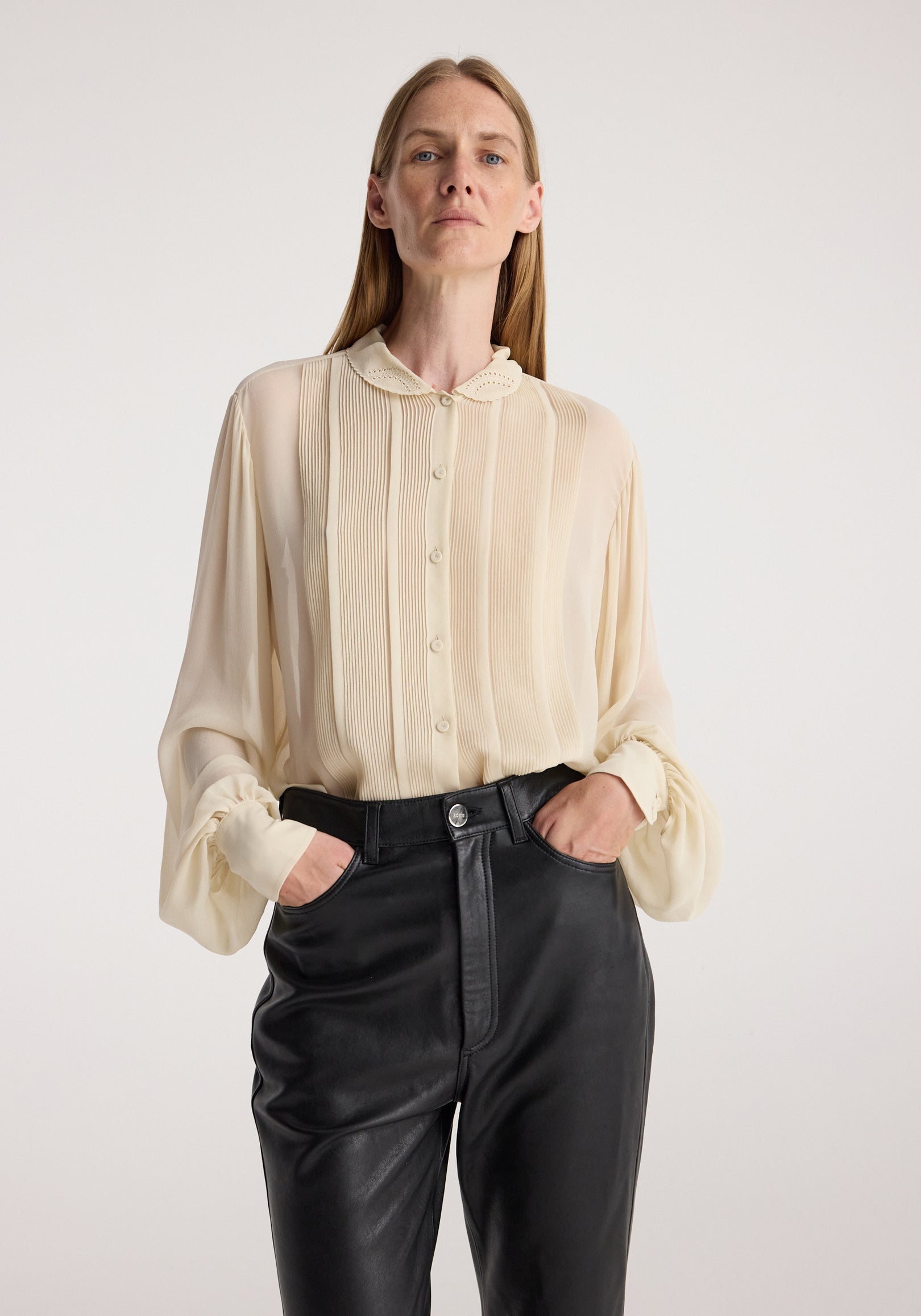 Woman wearing a cream button-up Romantic top | cream with pleated details and scalloped collar, paired with high-waisted black leather pants, posing with hands in pockets.