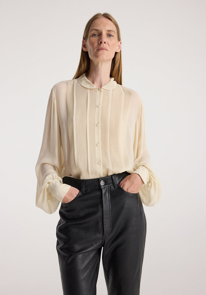 Woman wearing a cream button-up Romantic top | cream with pleated details and scalloped collar, paired with high-waisted black leather pants, posing with hands in pockets.