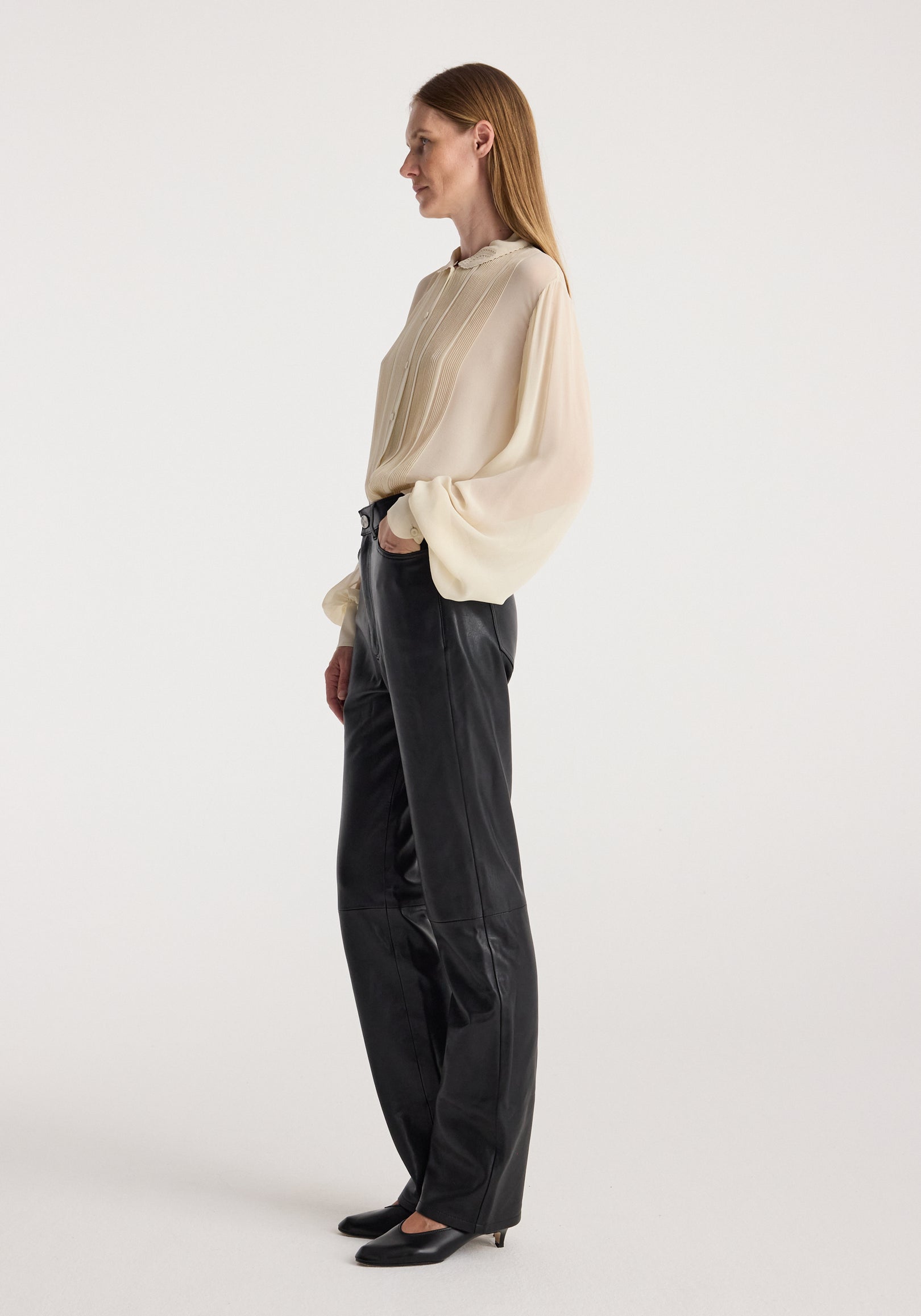 Side view of a woman wearing a cream colored Romantic top | cream with long, loose sleeves and pleated details, paired with black leather pants and black heels against a plain background.