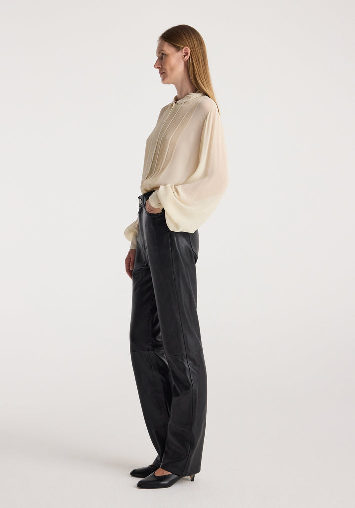 Side view of a woman wearing a cream colored Romantic top | cream with long, loose sleeves and pleated details, paired with black leather pants and black heels against a plain background.