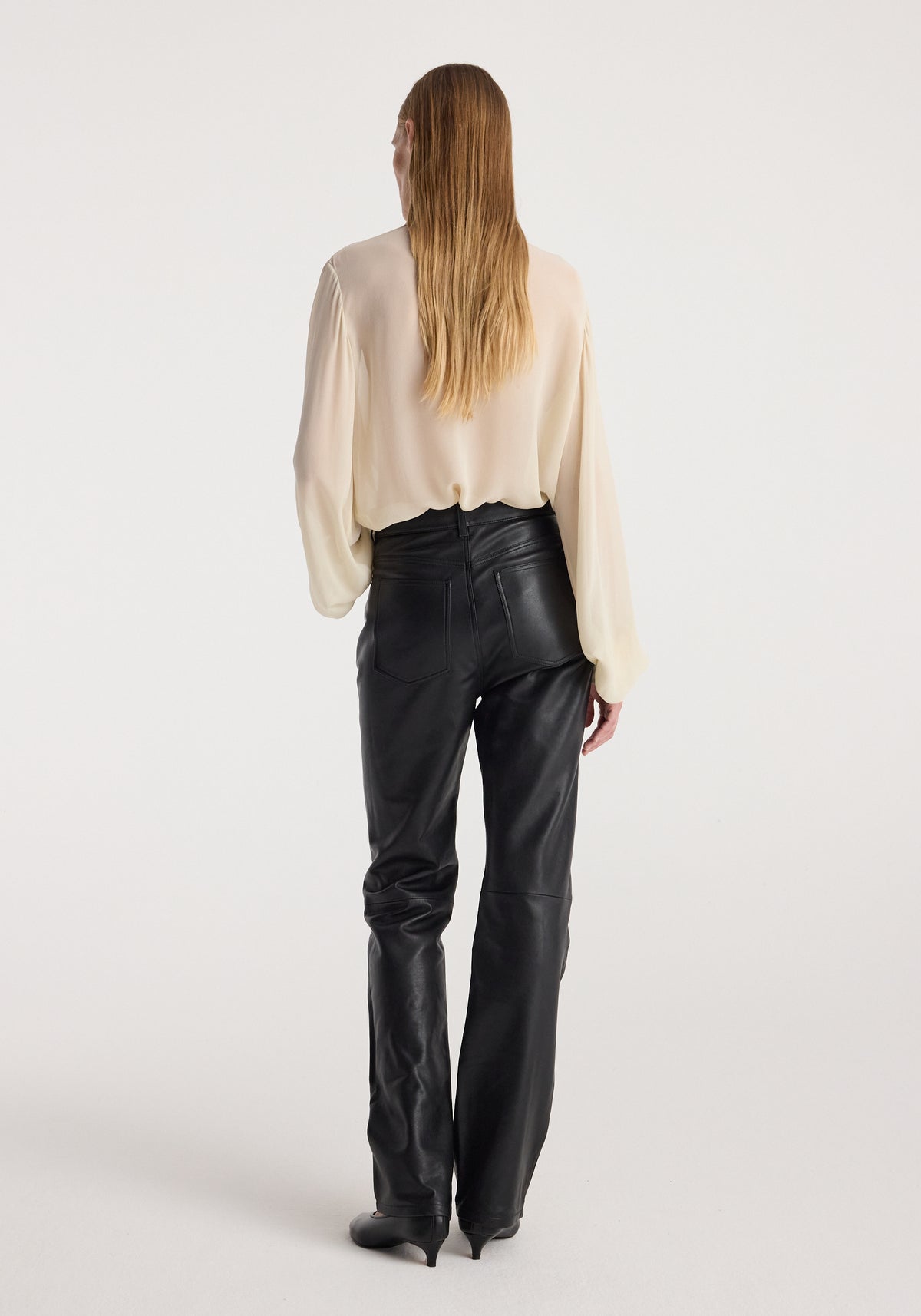 Woman with long blonde hair wearing a cream-colored Romantic top | cream with long sleeves, paired with high-waisted black leather pants, standing against a plain background viewed from behind.