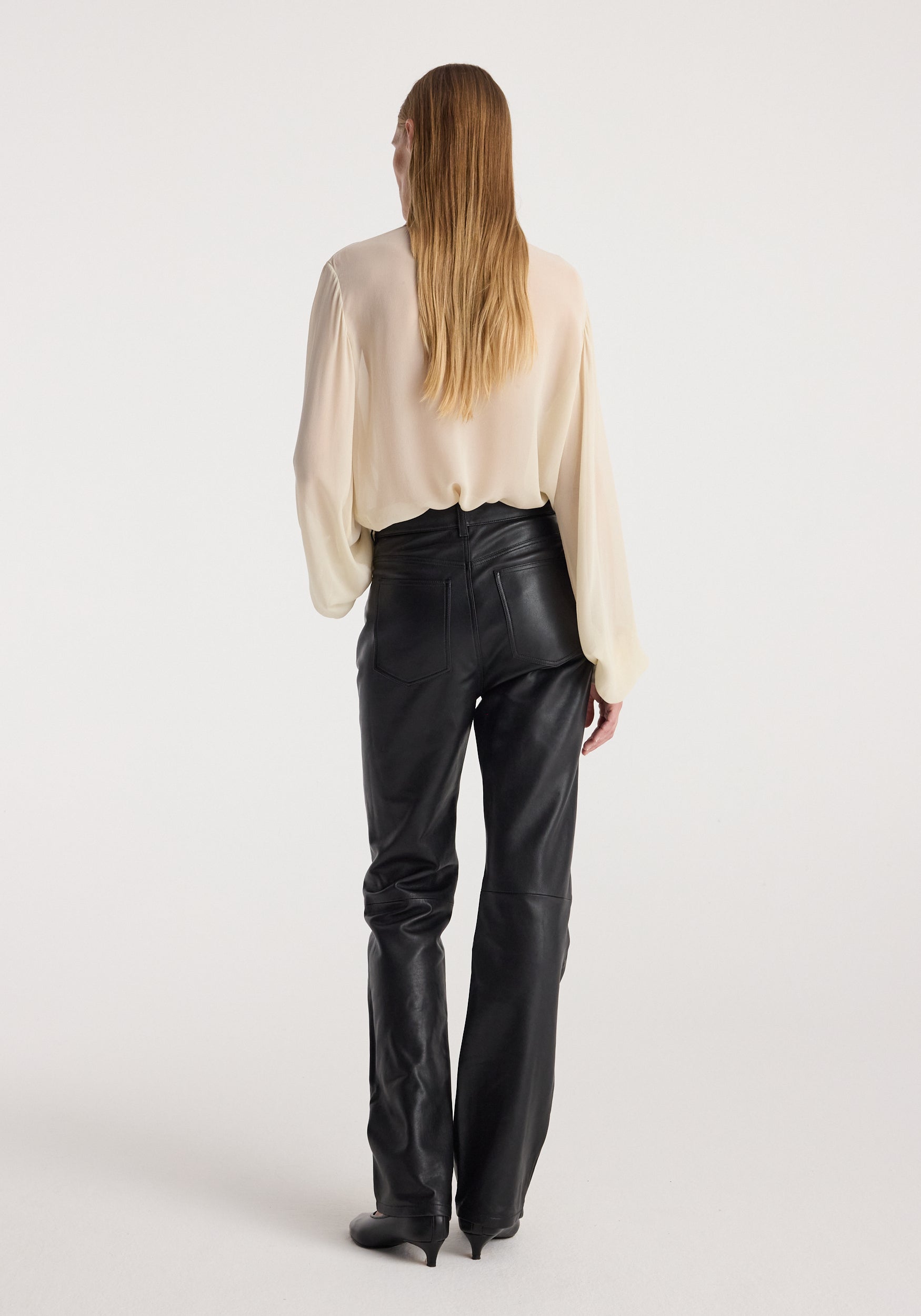 Woman with long blonde hair wearing a cream-colored Romantic top | cream with long sleeves, paired with high-waisted black leather pants, standing against a plain background viewed from behind.