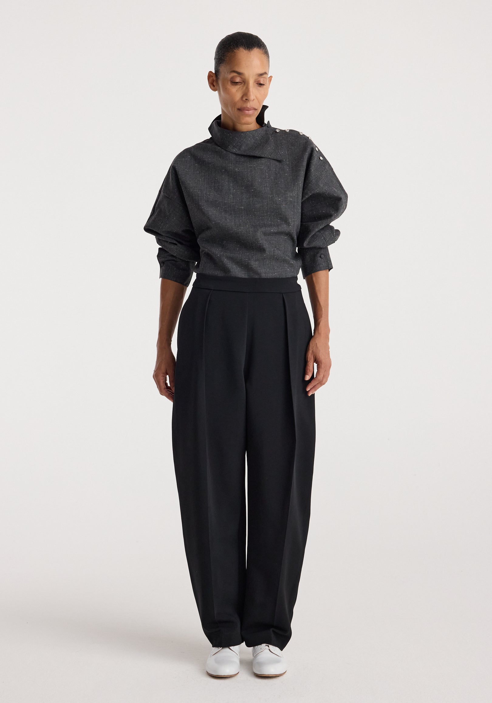 Woman wearing a grey slub textured top with a high, asymmetrical collar and button details on the shoulder, paired with loose black trousers and white shoes, styled in the Reimagined collar top | grey slub.