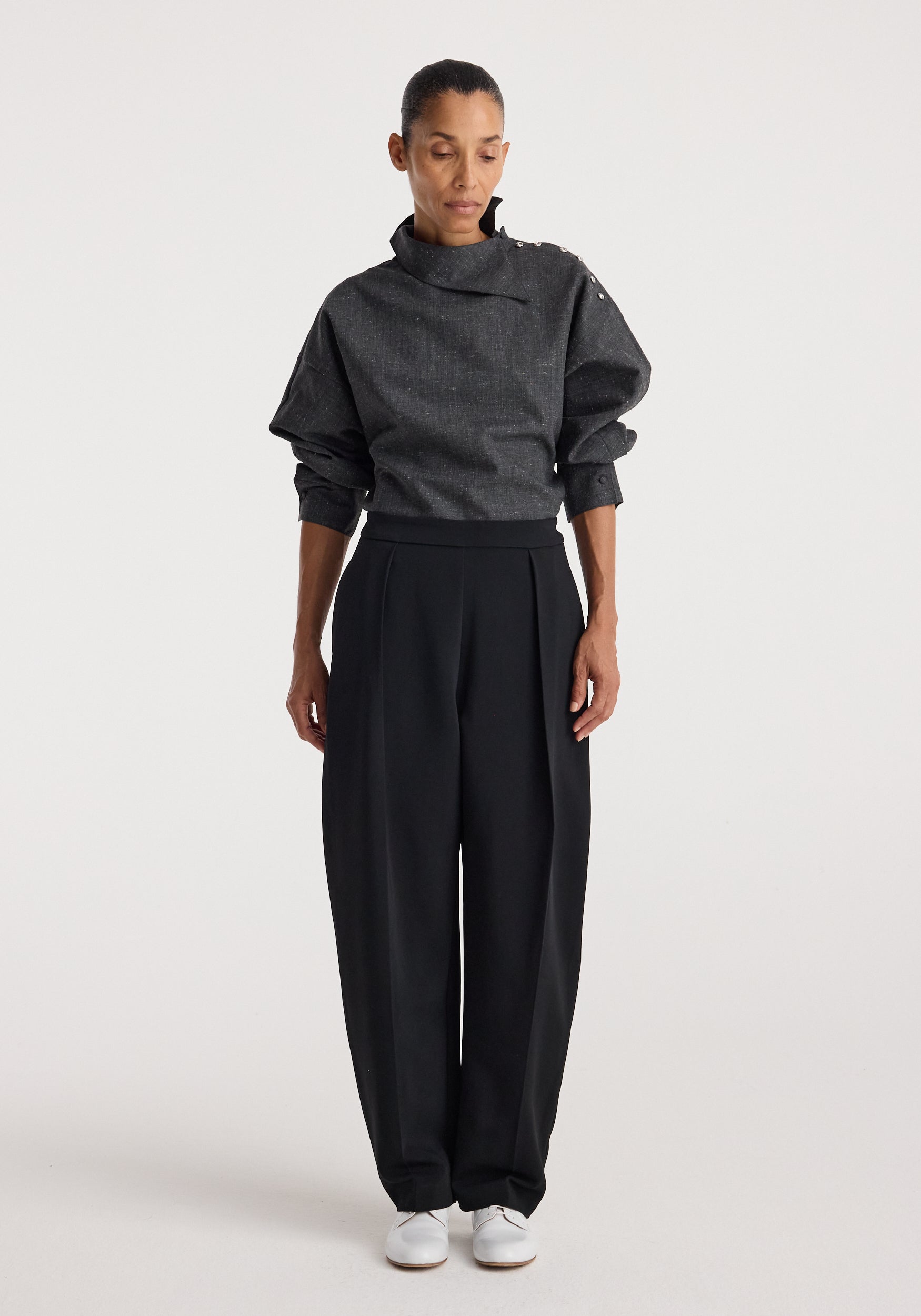 Woman wearing a grey slub textured top with a high, asymmetrical collar and button details on the shoulder, paired with loose black trousers and white shoes, styled in the Reimagined collar top | grey slub.