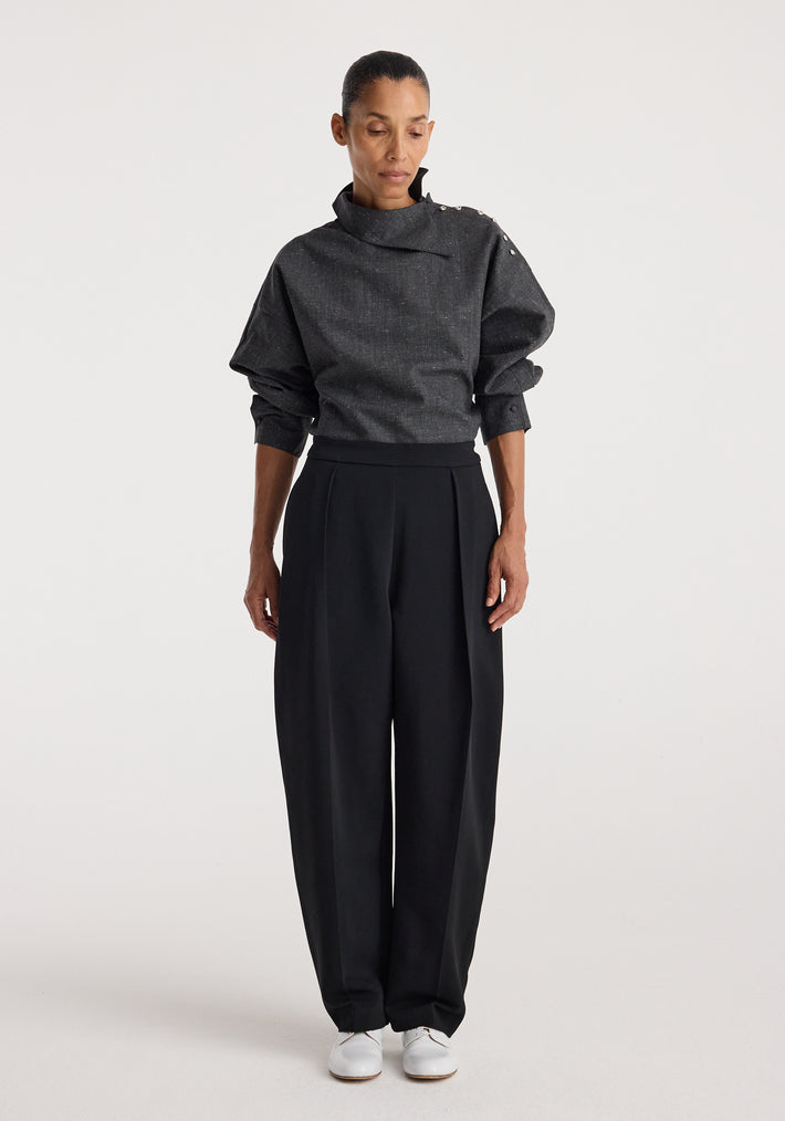 Woman wearing a grey slub textured top with a high, asymmetrical collar and button details on the shoulder, paired with loose black trousers and white shoes, styled in the Reimagined collar top | grey slub.