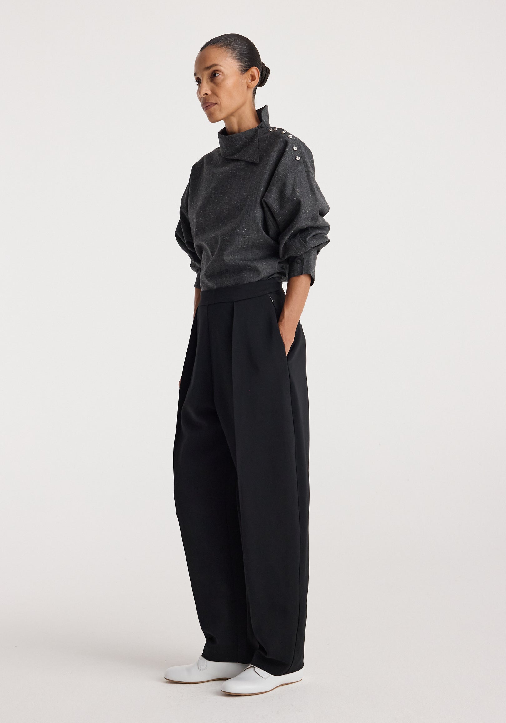 Woman wearing a dark grey slub fabric top with a high collar and button detail on the shoulder, styled with loose black pants and white shoes, Reimagined collar top | grey slub.