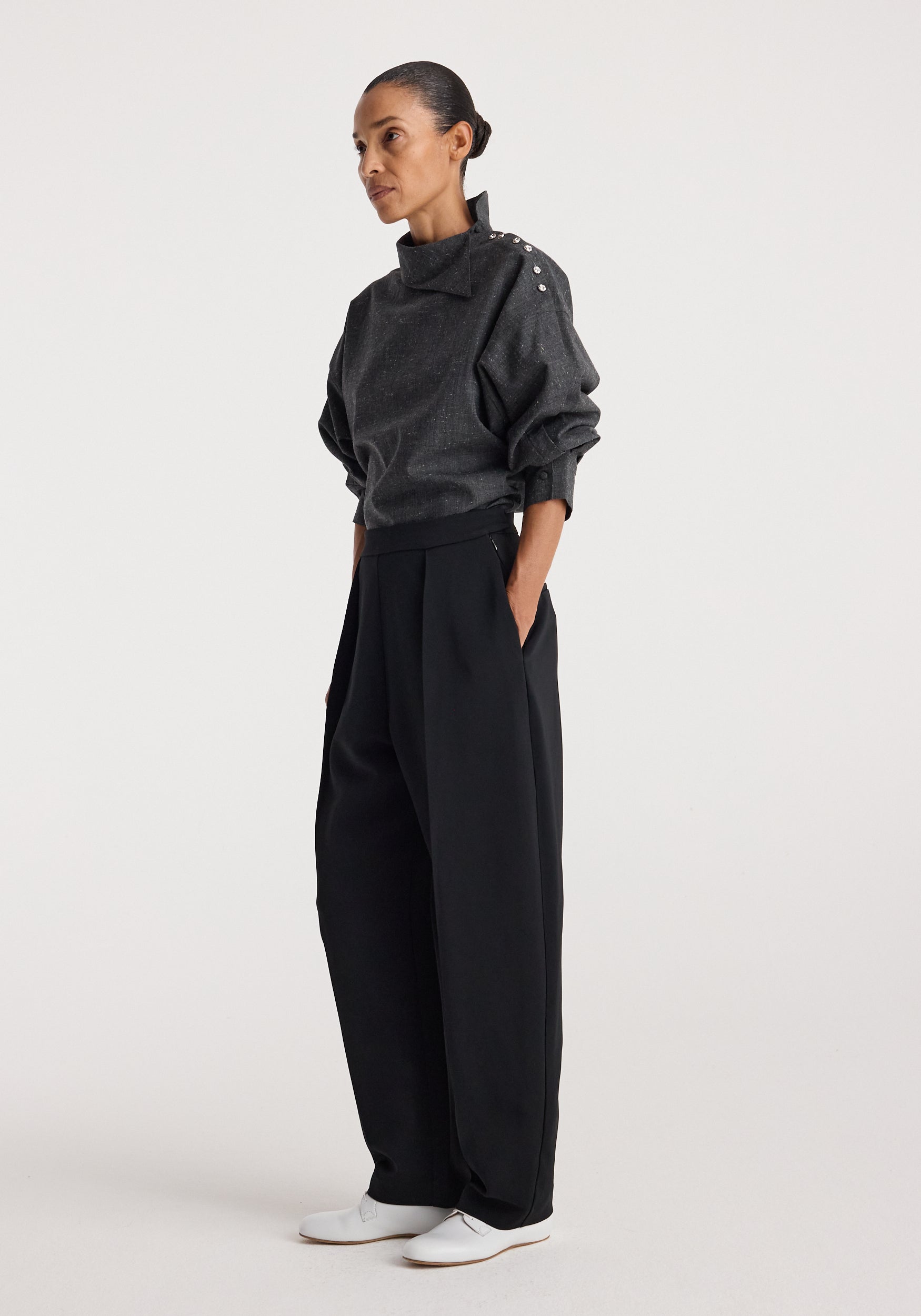 Woman wearing a dark grey slub fabric top with a high collar and button detail on the shoulder, styled with loose black pants and white shoes, Reimagined collar top | grey slub.