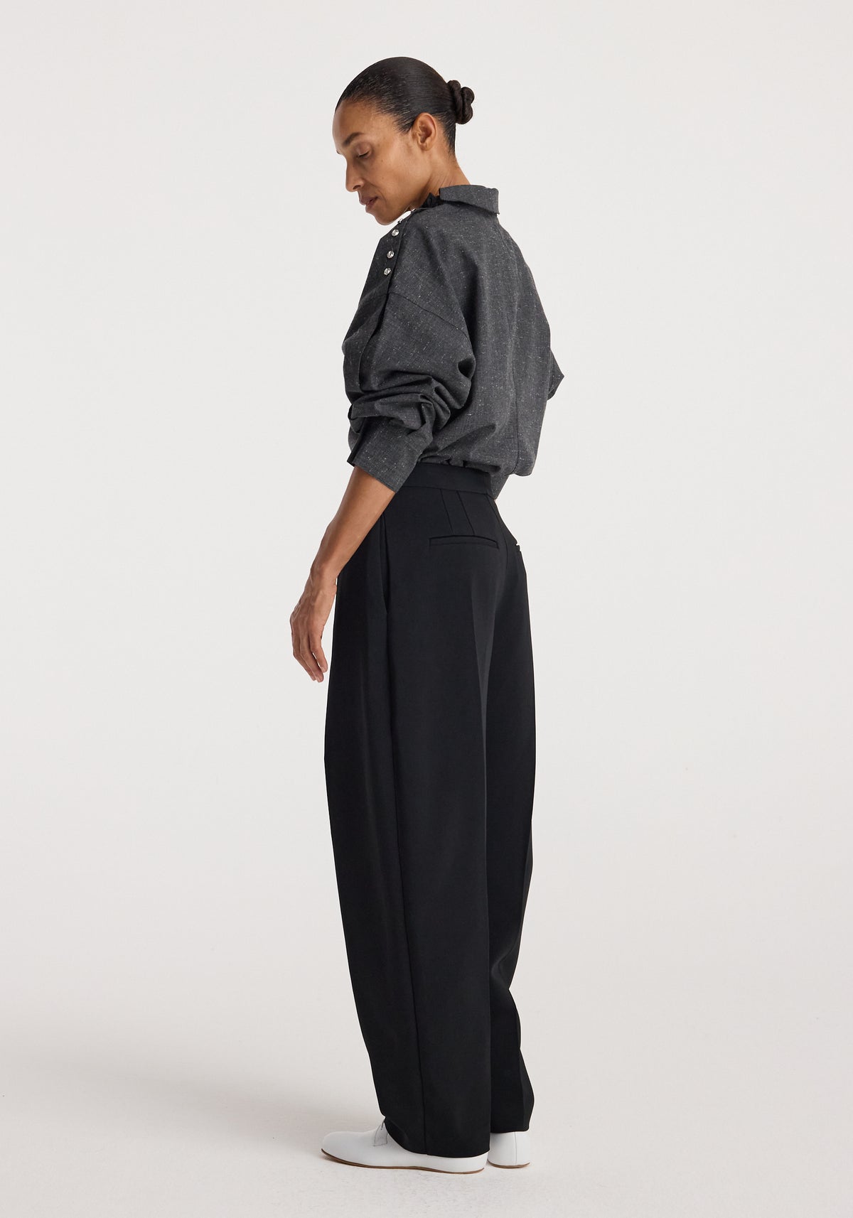 Side view of a woman wearing a grey slub textured Reimagined collar top with long sleeves and button details on the shoulder, paired with black wide-leg pants and white shoes.