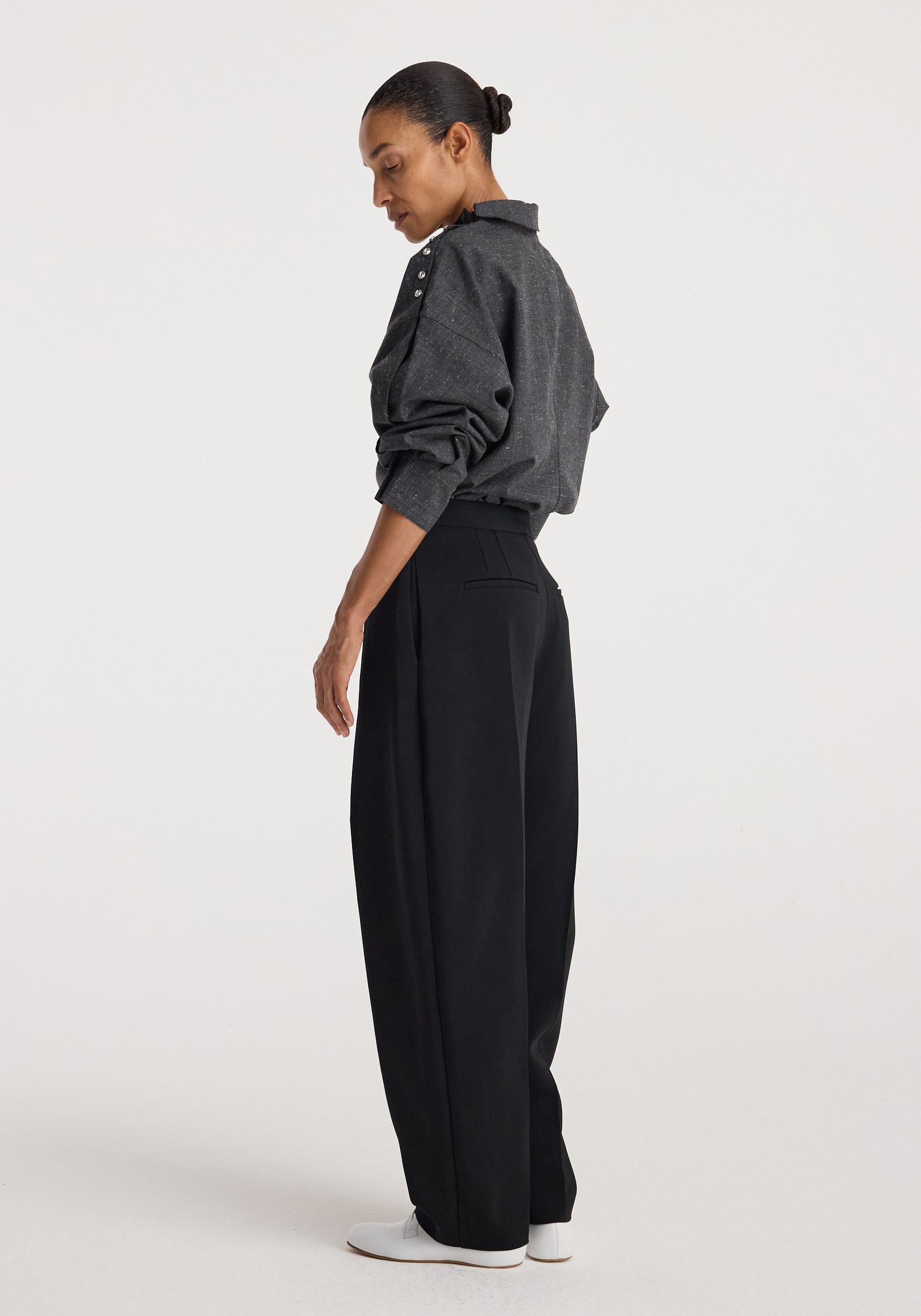 Side view of a woman wearing a grey slub textured Reimagined collar top with long sleeves and button details on the shoulder, paired with black wide-leg pants and white shoes.