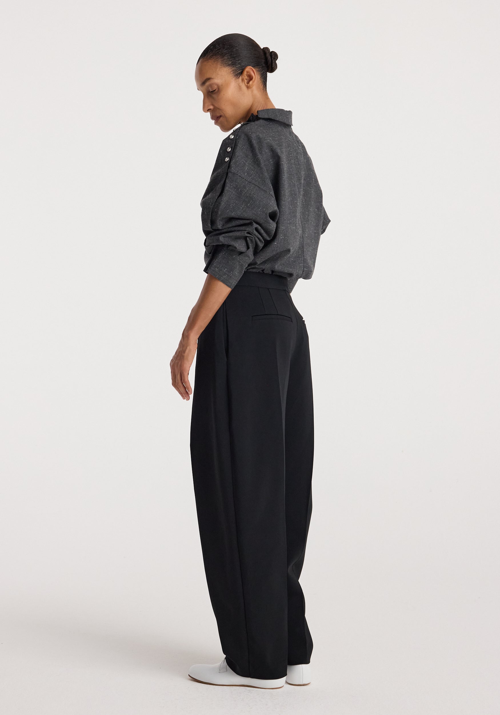 Side view of a woman wearing a grey slub textured Reimagined collar top with long sleeves and button details on the shoulder, paired with black wide-leg pants and white shoes.
