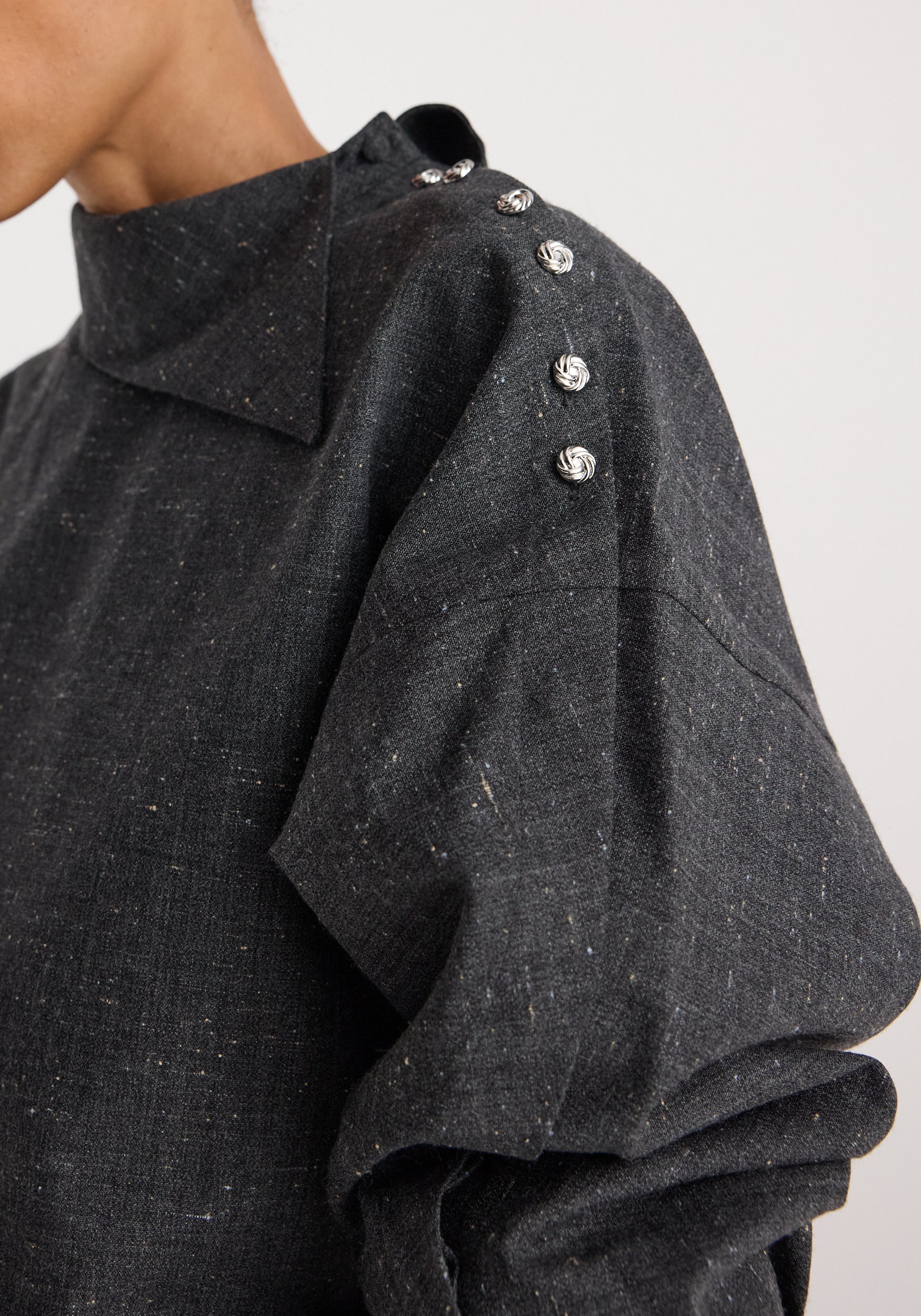 Close-up of a person wearing a Reimagined collar top | grey slub featuring a high collar and silver decorative buttons along the shoulder seam on textured grey fabric.