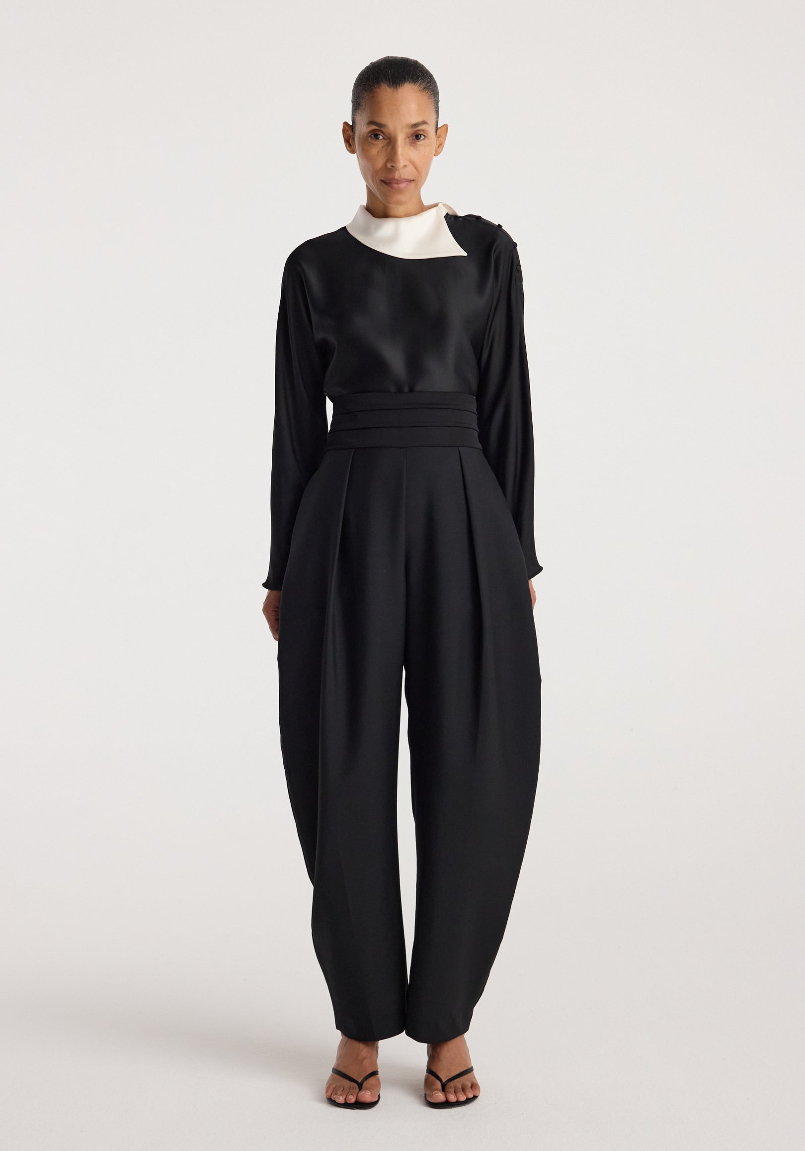 Woman wearing a black top with a contrasting white reimagined collar and long sleeves paired with high-waisted, pleated black pants and black sandals, featuring the Reimagined collar top | black.