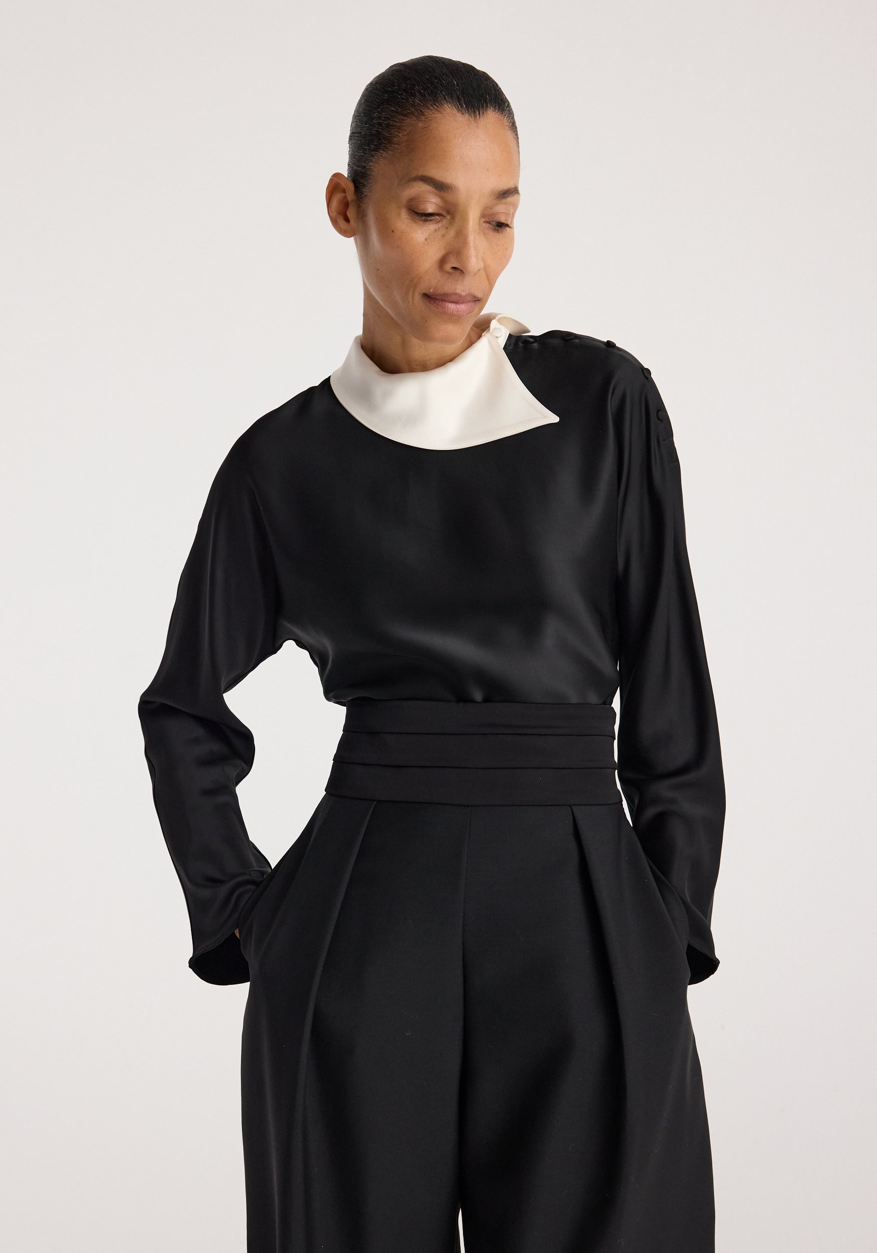 Woman wearing a black garment with a white asymmetrical collar and long sleeves, styled with high-waisted black pants, showcasing the Reimagined collar top | black.