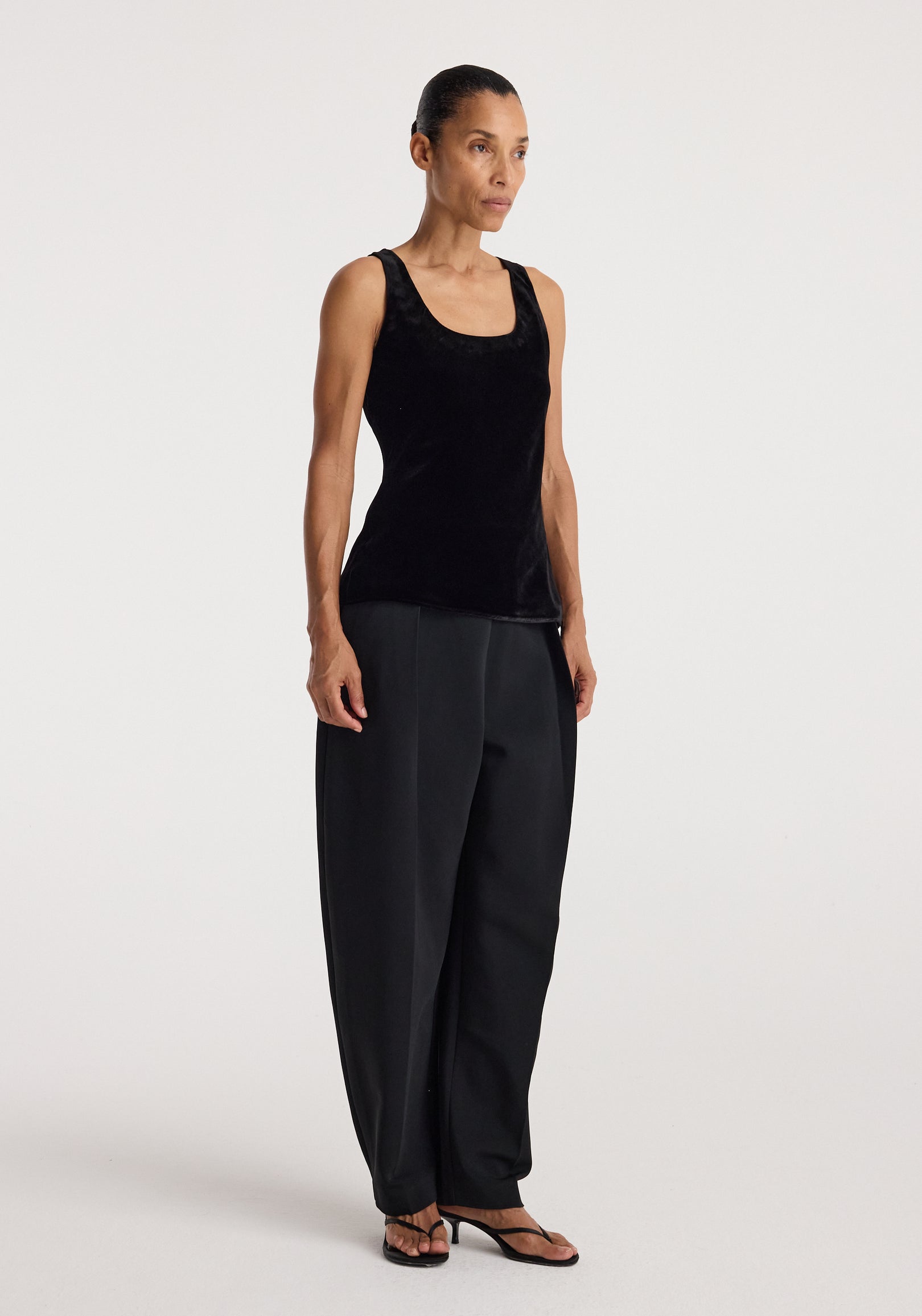 Woman wearing a black scoop-neck silk velvet top | black paired with loose black trousers and black sandals, standing against a plain light background.