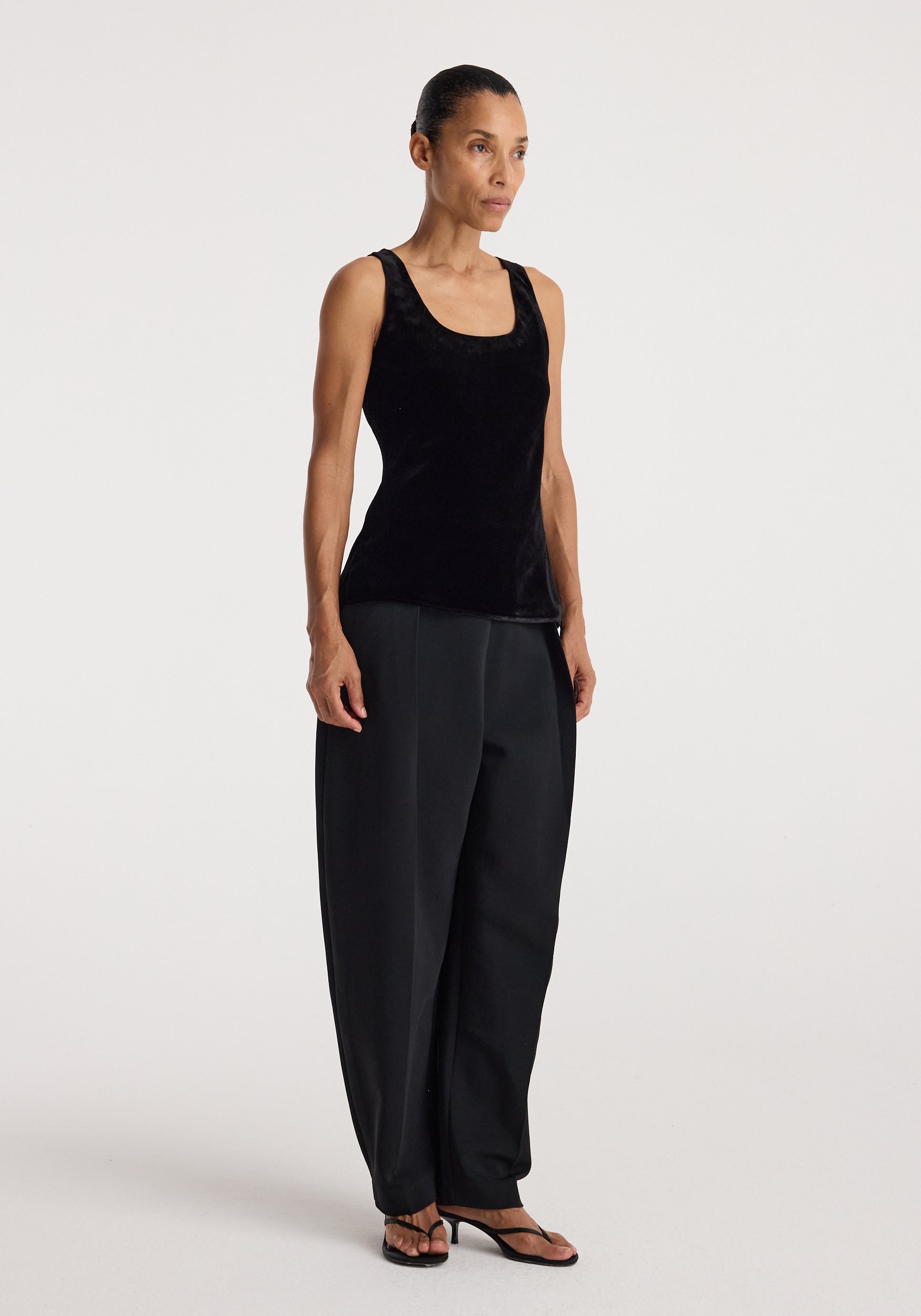 Woman wearing a black scoop-neck silk velvet top | black paired with loose black trousers and black sandals, standing against a plain light background.