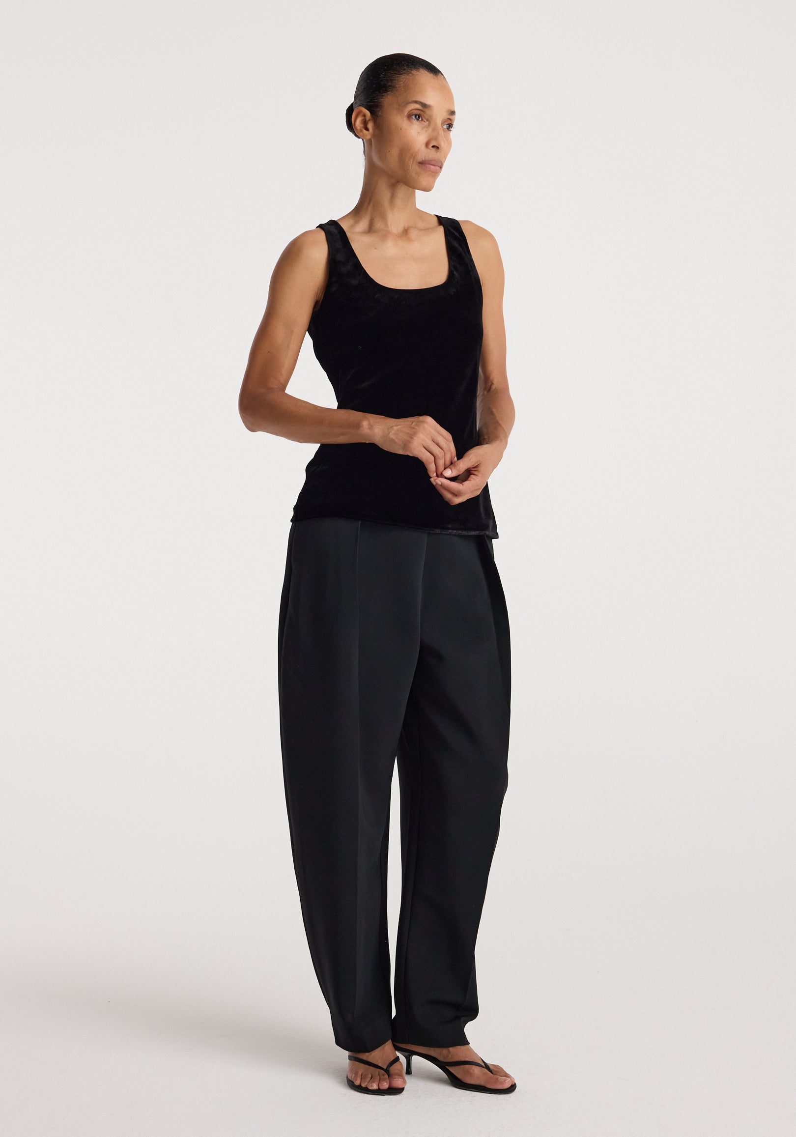 Woman wearing a black Scoop-neck silk velvet top | black paired with loose black pants and black sandals, standing against a plain white background.