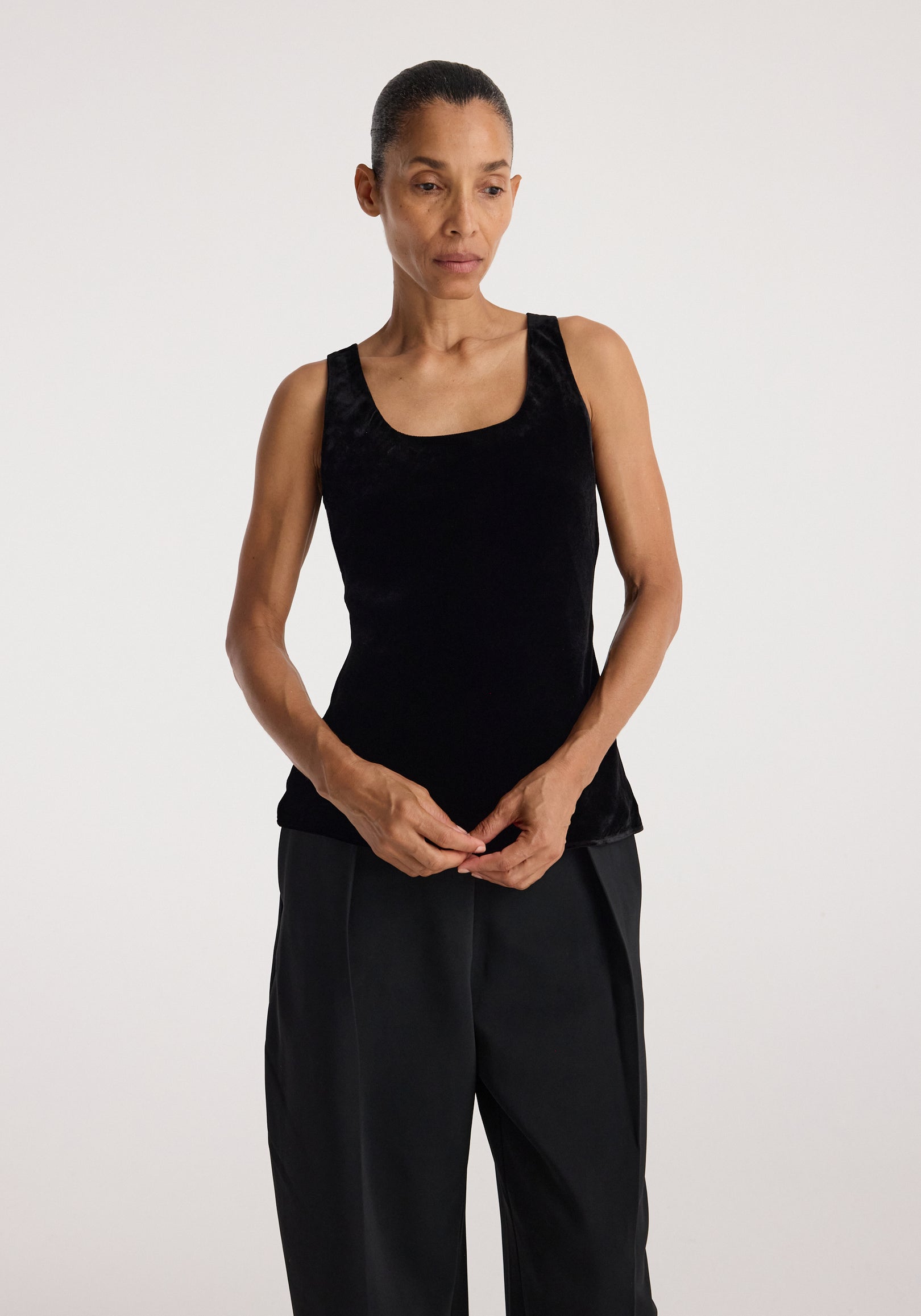 Woman wearing a sleeveless black scoop-neck silk velvet top | black paired with loose black pants against a plain light background.