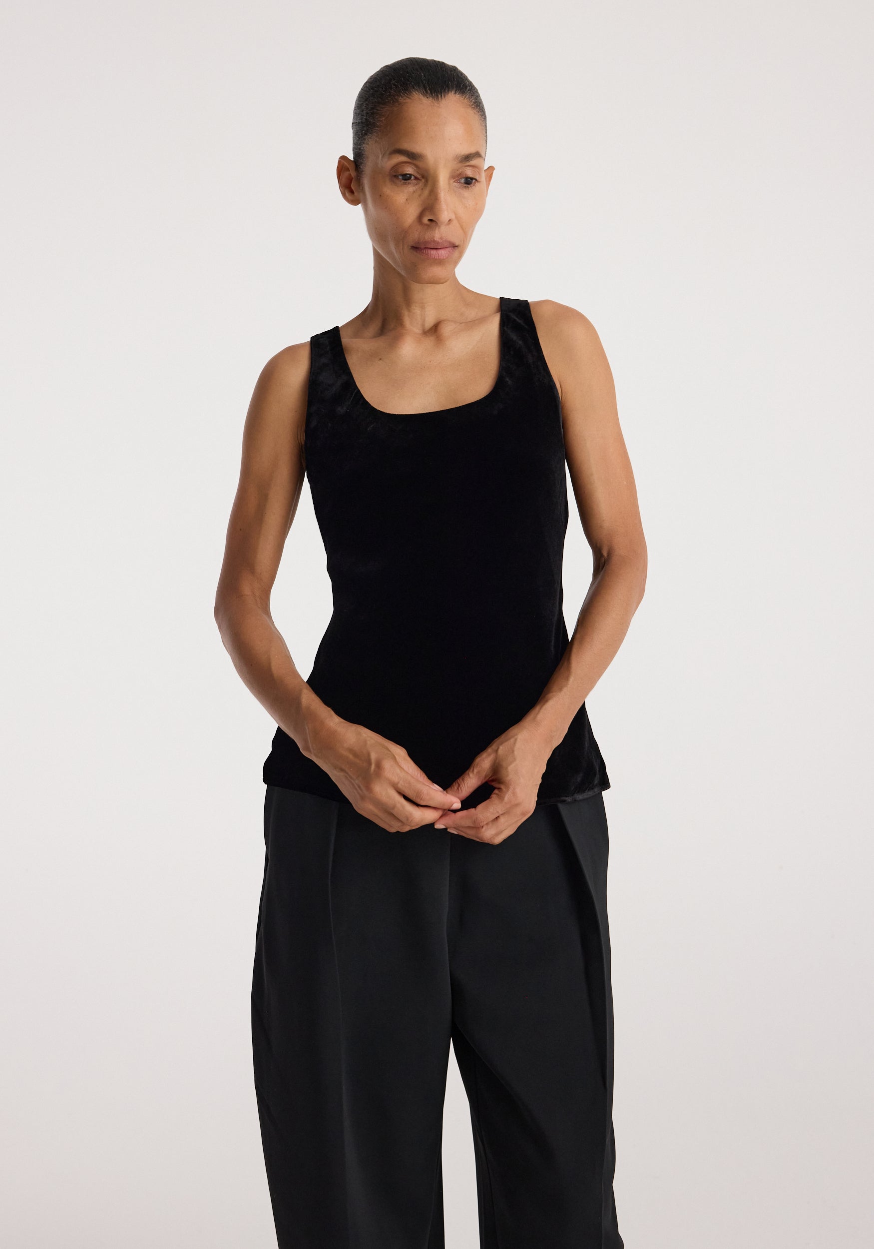 Woman wearing a sleeveless black scoop-neck silk velvet top | black paired with loose black pants against a plain light background.