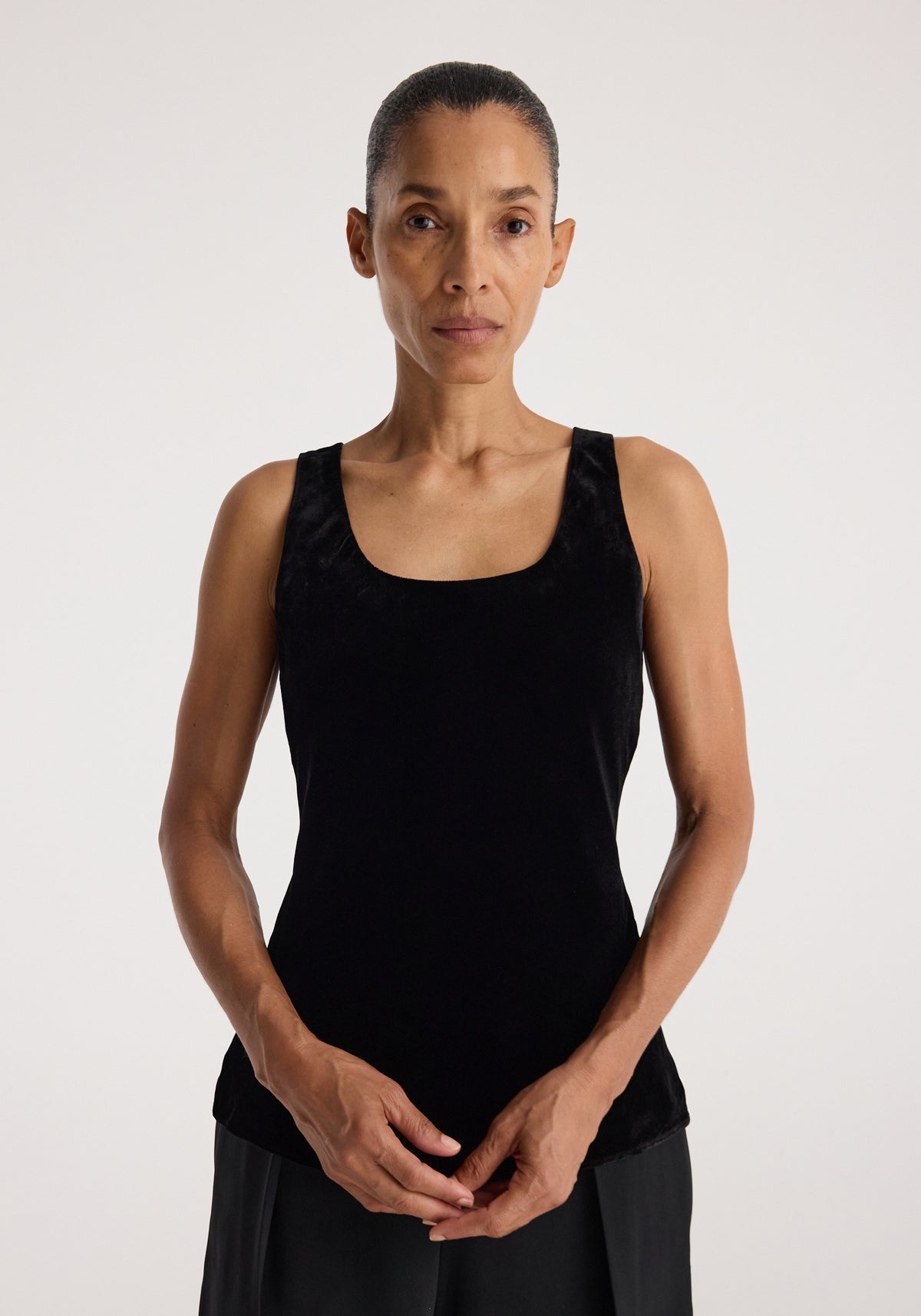 Woman wearing a sleeveless scoop-neck silk velvet top | black, standing against a plain light background with hands gently clasped in front.