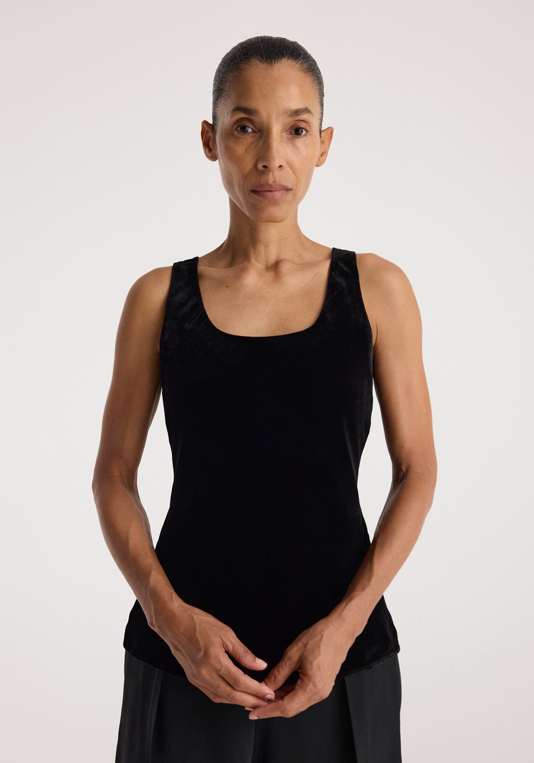 Woman wearing a sleeveless scoop-neck silk velvet top | black, standing against a plain light background with hands gently clasped in front.