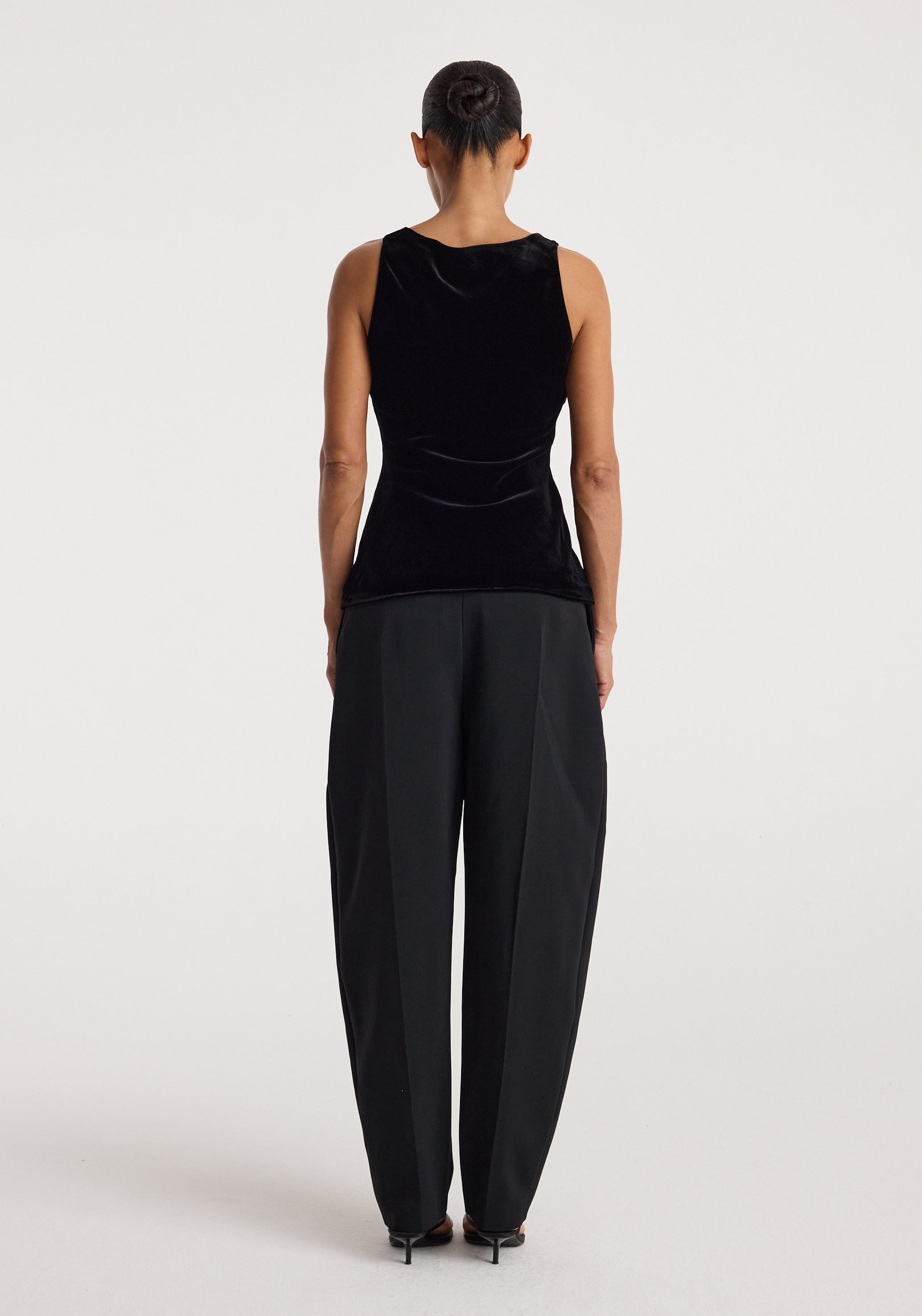 Woman wearing a black scoop-neck silk velvet top | black, shown from the back, paired with loose-fitting black trousers and black heels against a plain background.
