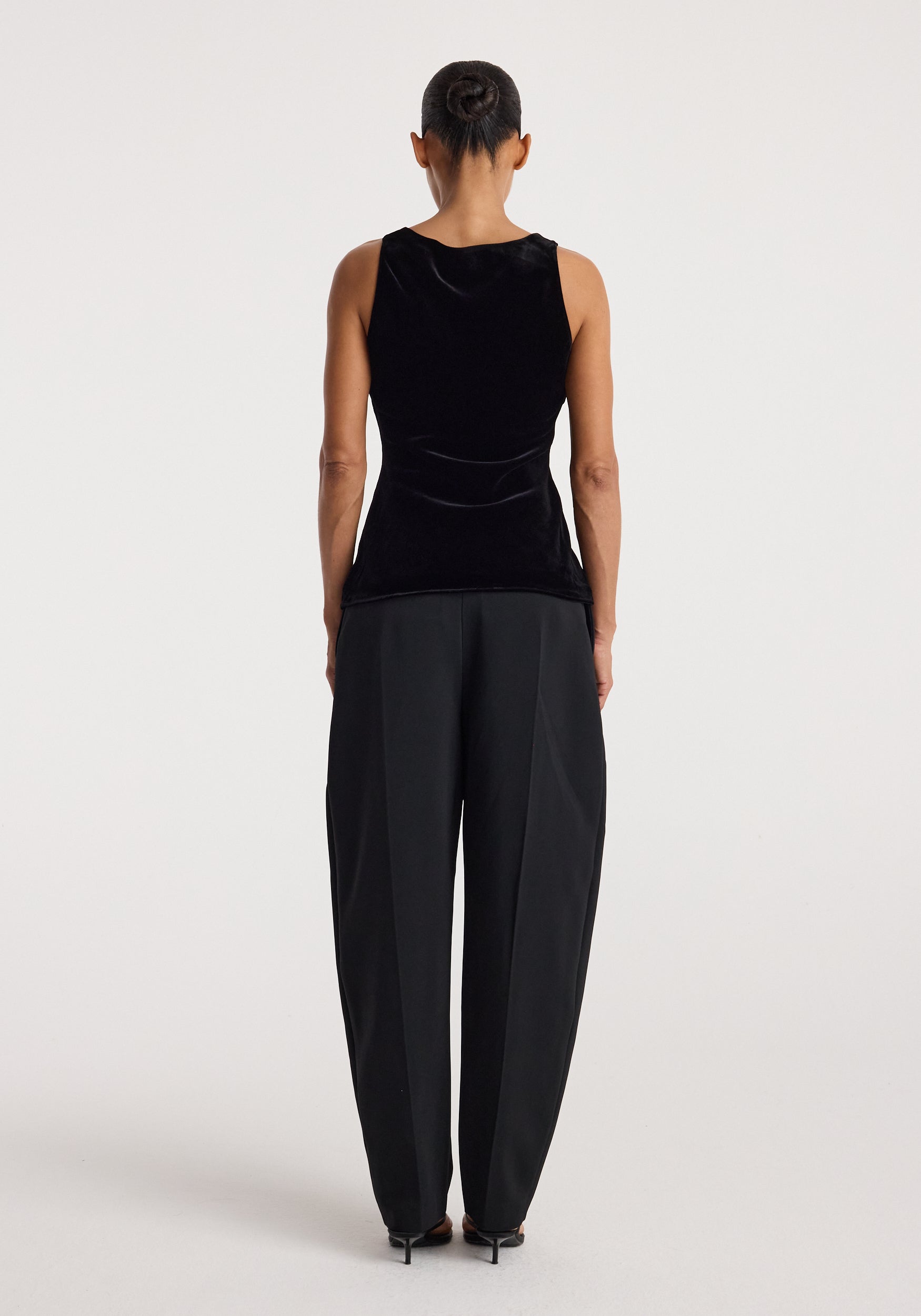 Woman wearing a black scoop-neck silk velvet top | black, shown from the back, paired with loose-fitting black trousers and black heels against a plain background.