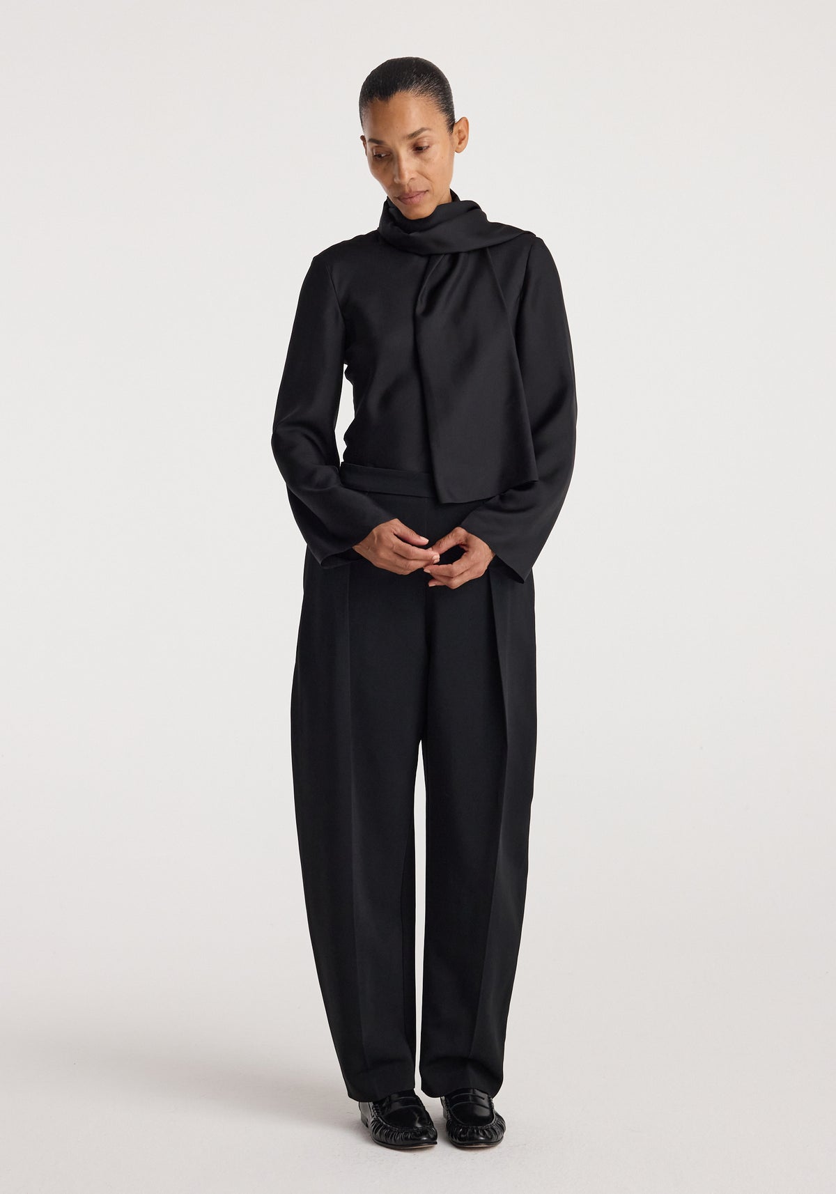 Woman wearing a black Silk foulard top | black with a scarf-like neckline, paired with loose black trousers and black shoes, standing against a plain background.