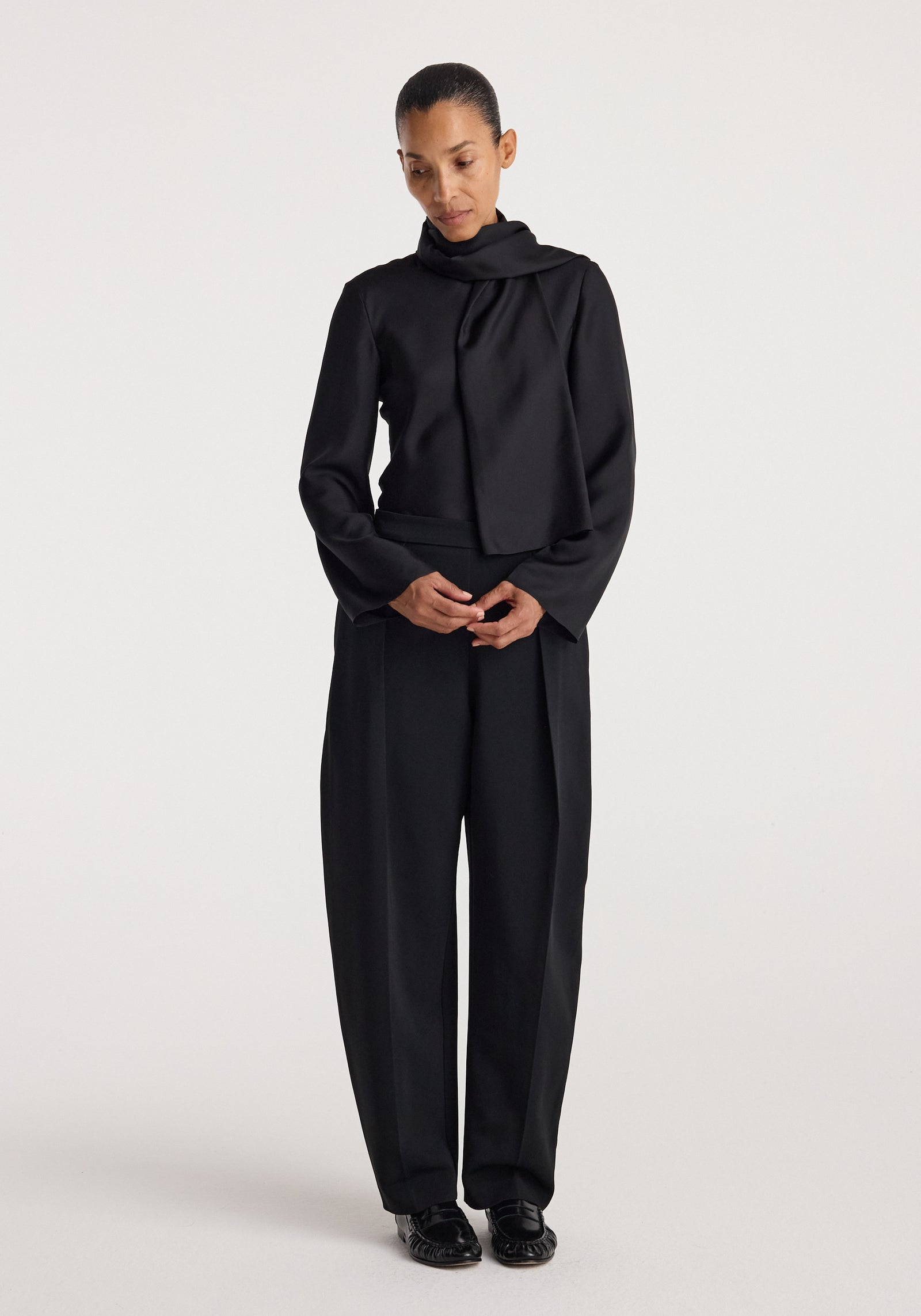 Woman wearing a black Silk foulard top | black with a scarf-like neckline, paired with loose black trousers and black shoes, standing against a plain background.