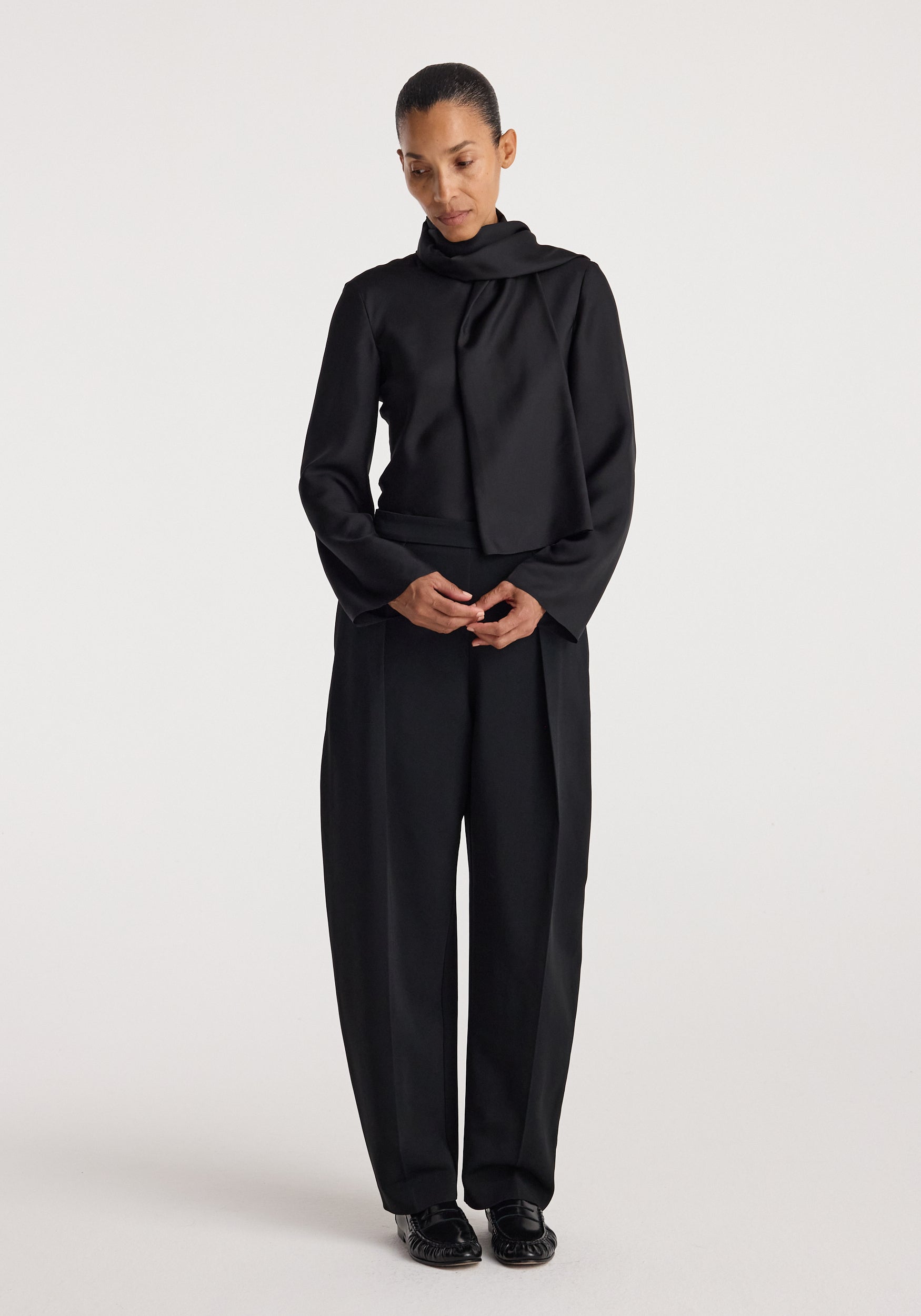 Woman wearing a black Silk foulard top | black with a scarf-like neckline, paired with loose black trousers and black shoes, standing against a plain background.