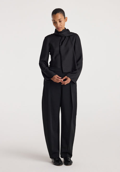 Woman wearing a black Silk foulard top | black with a scarf-like neckline, paired with loose black trousers and black shoes, standing against a plain background.