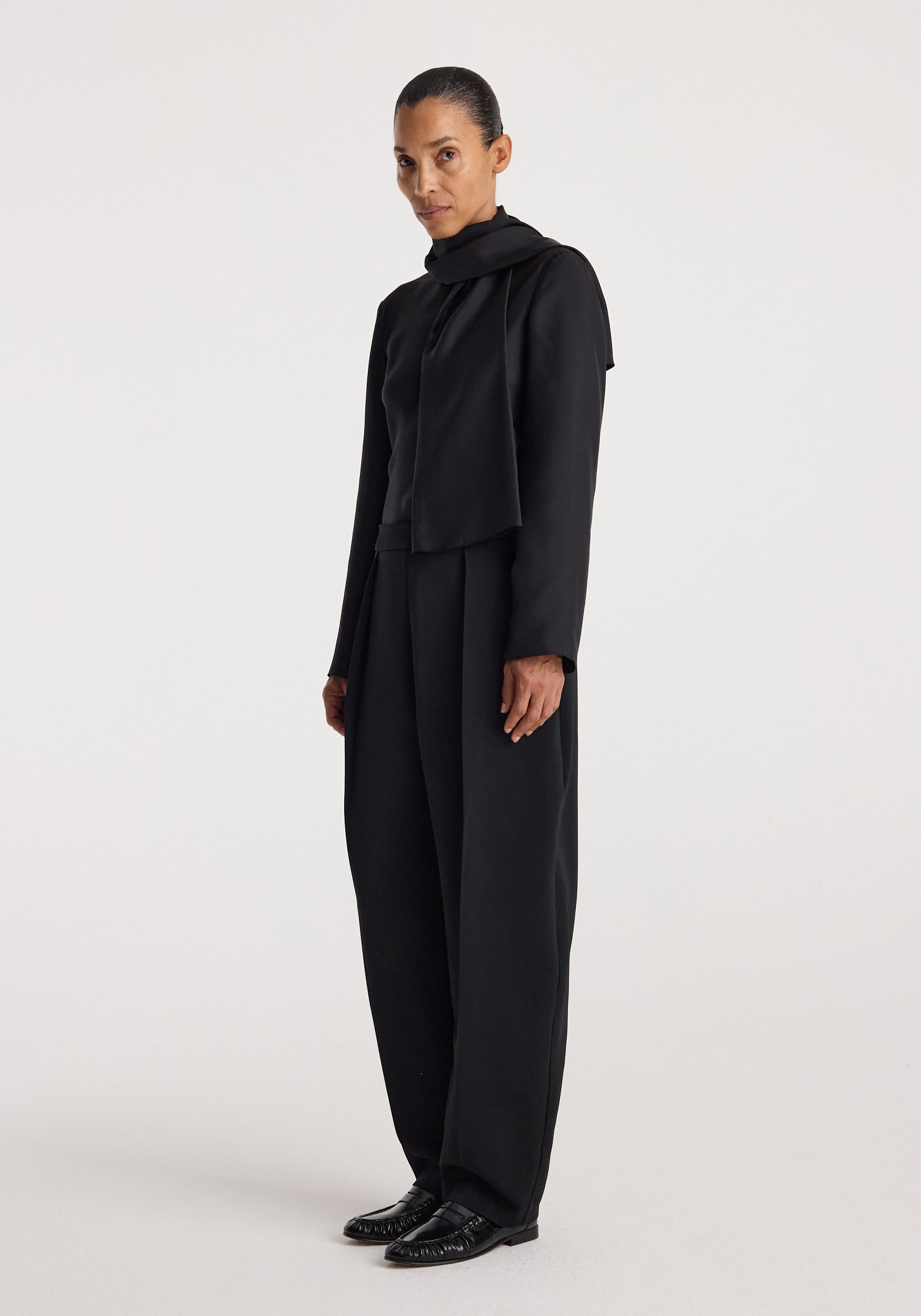 Woman wearing a black silk foulard top | black with long sleeves and a high neckline, paired with matching black wide-leg trousers and black shoes against a plain background.