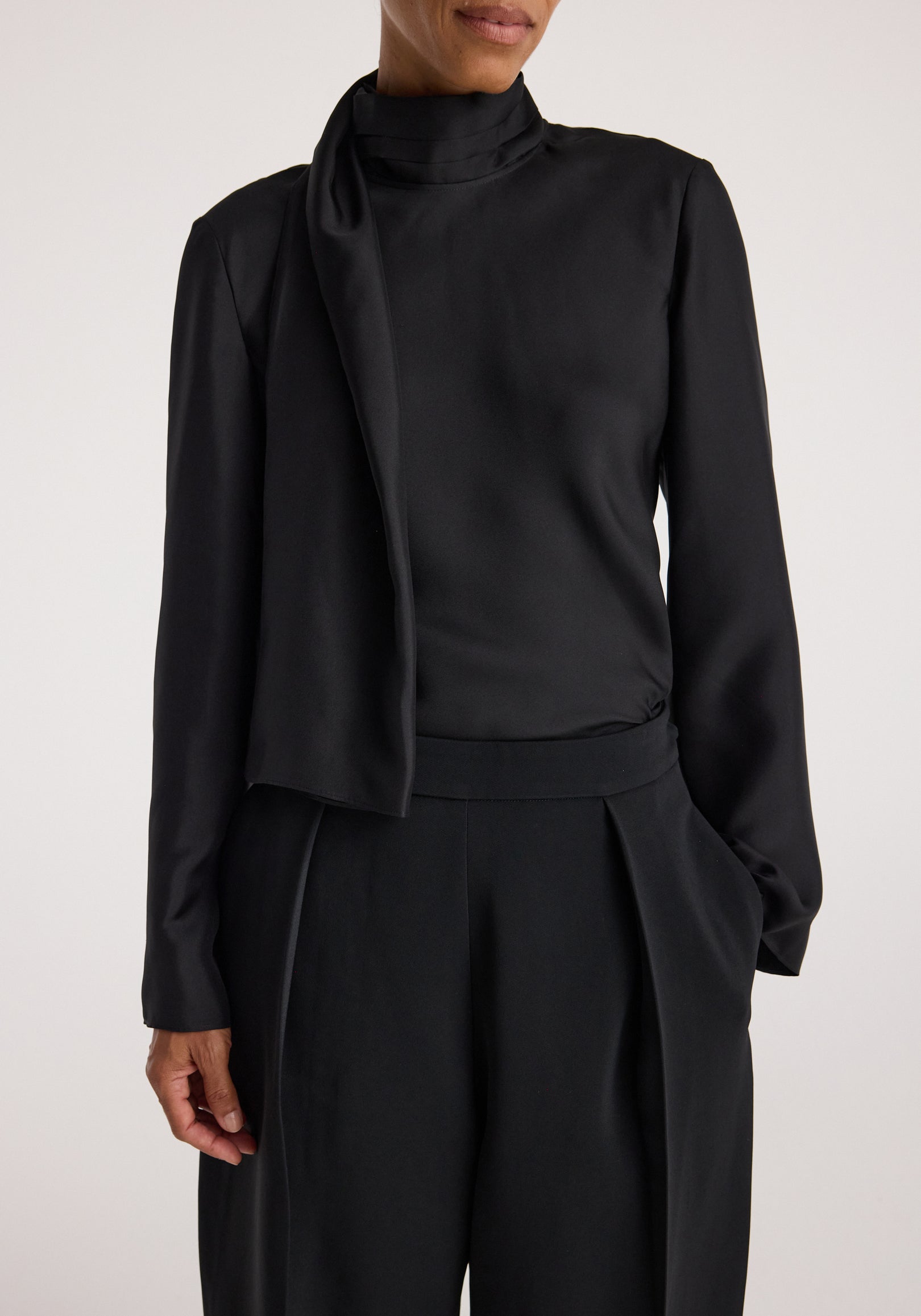 Woman wearing a sleek black Silk foulard top | black featuring a high neck with an attached scarf detail and long sleeves paired with black high-waisted pants.