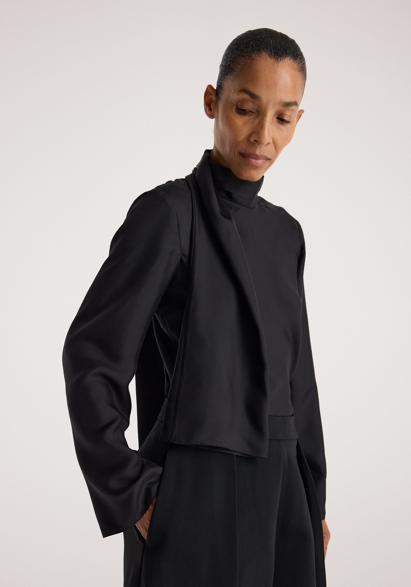 Woman wearing a black silk foulard top | black with long sleeves and a high neckline, shown from the waist up against a plain background.