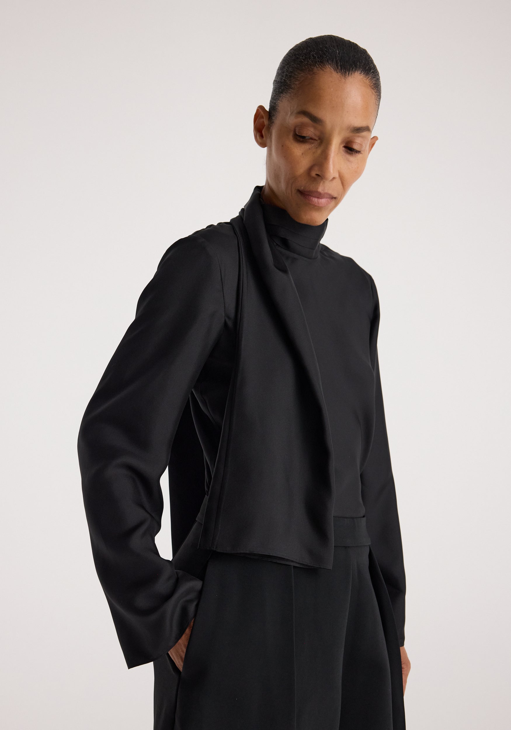 Woman wearing a black silk foulard top | black with long sleeves and a high neckline, shown from the waist up against a plain background.