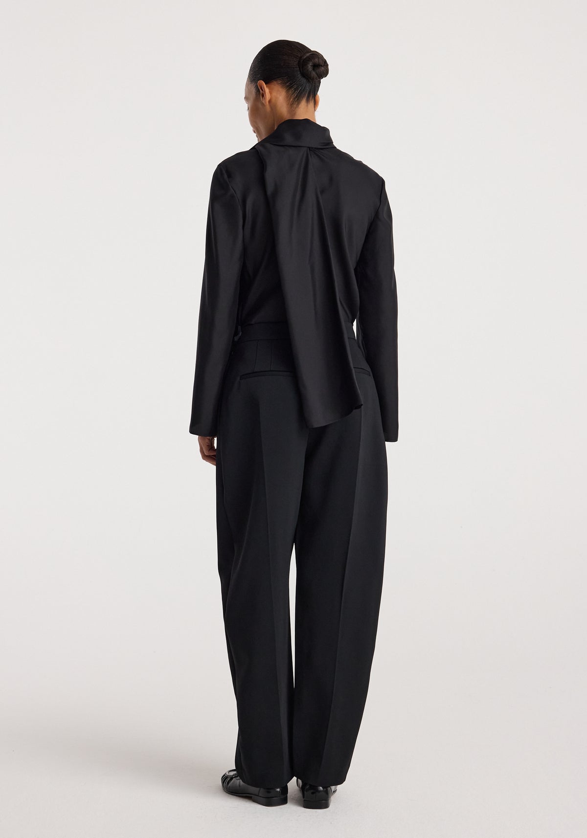 Woman seen from back wearing a black Silk foulard top | black with long sleeves and a draped detail, paired with wide-leg black trousers and black shoes.