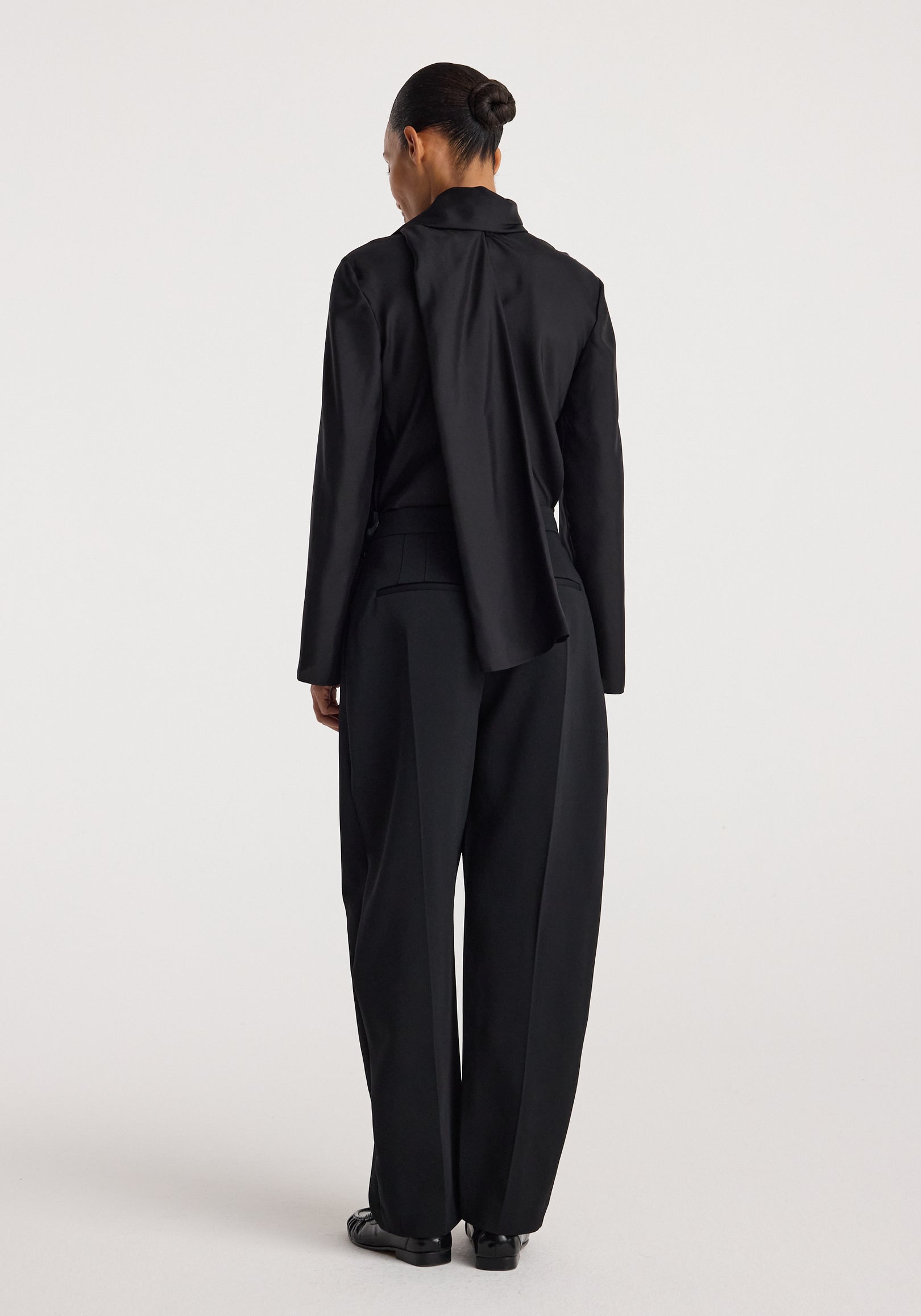 Woman seen from back wearing a black Silk foulard top | black with long sleeves and a draped detail, paired with wide-leg black trousers and black shoes.