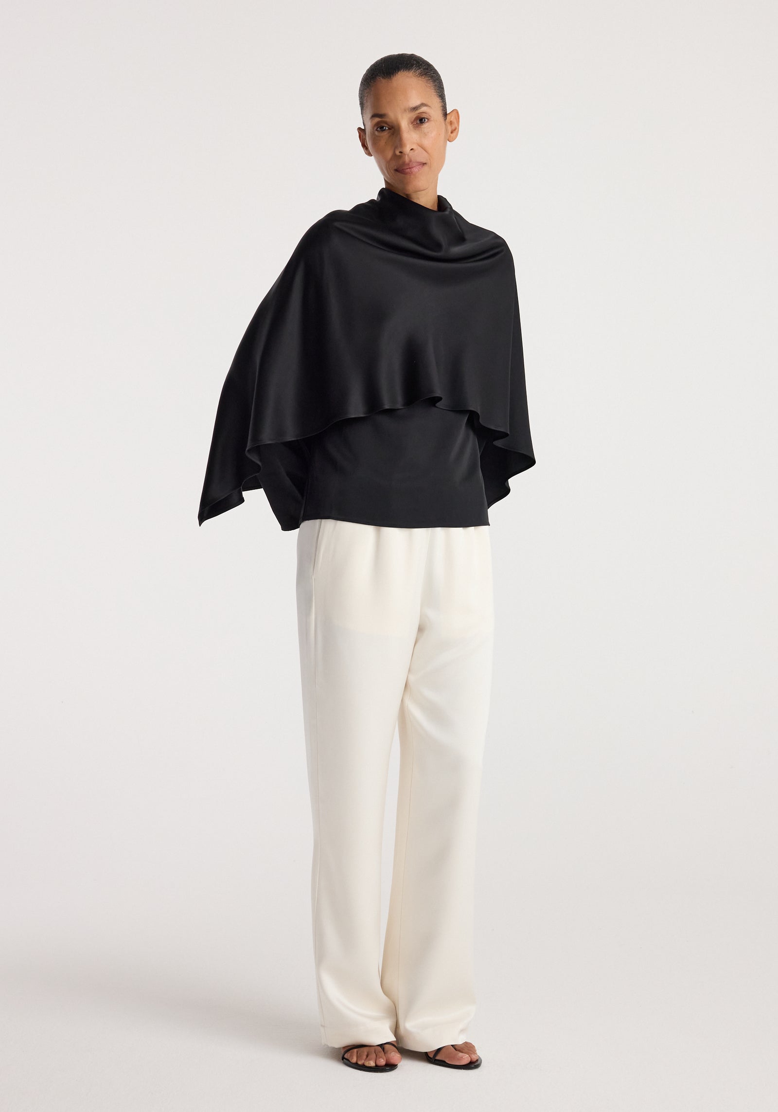 Woman wearing a black Satin cape top | black with a draped silhouette paired with loose white trousers and black open-toe sandals against a plain background.