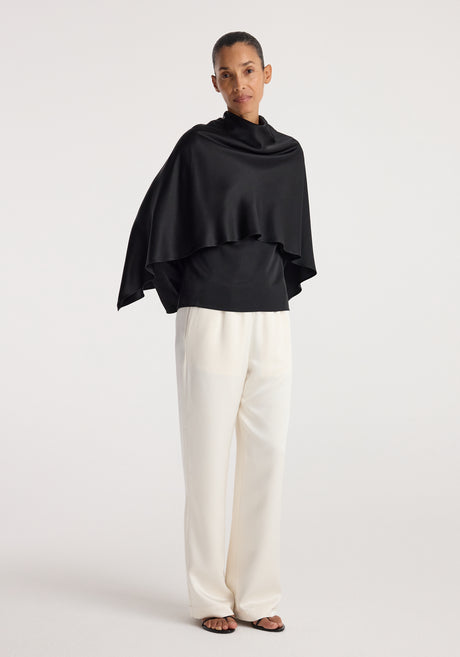 Woman wearing a black Satin cape top | black with a draped silhouette paired with loose white trousers and black open-toe sandals against a plain background.