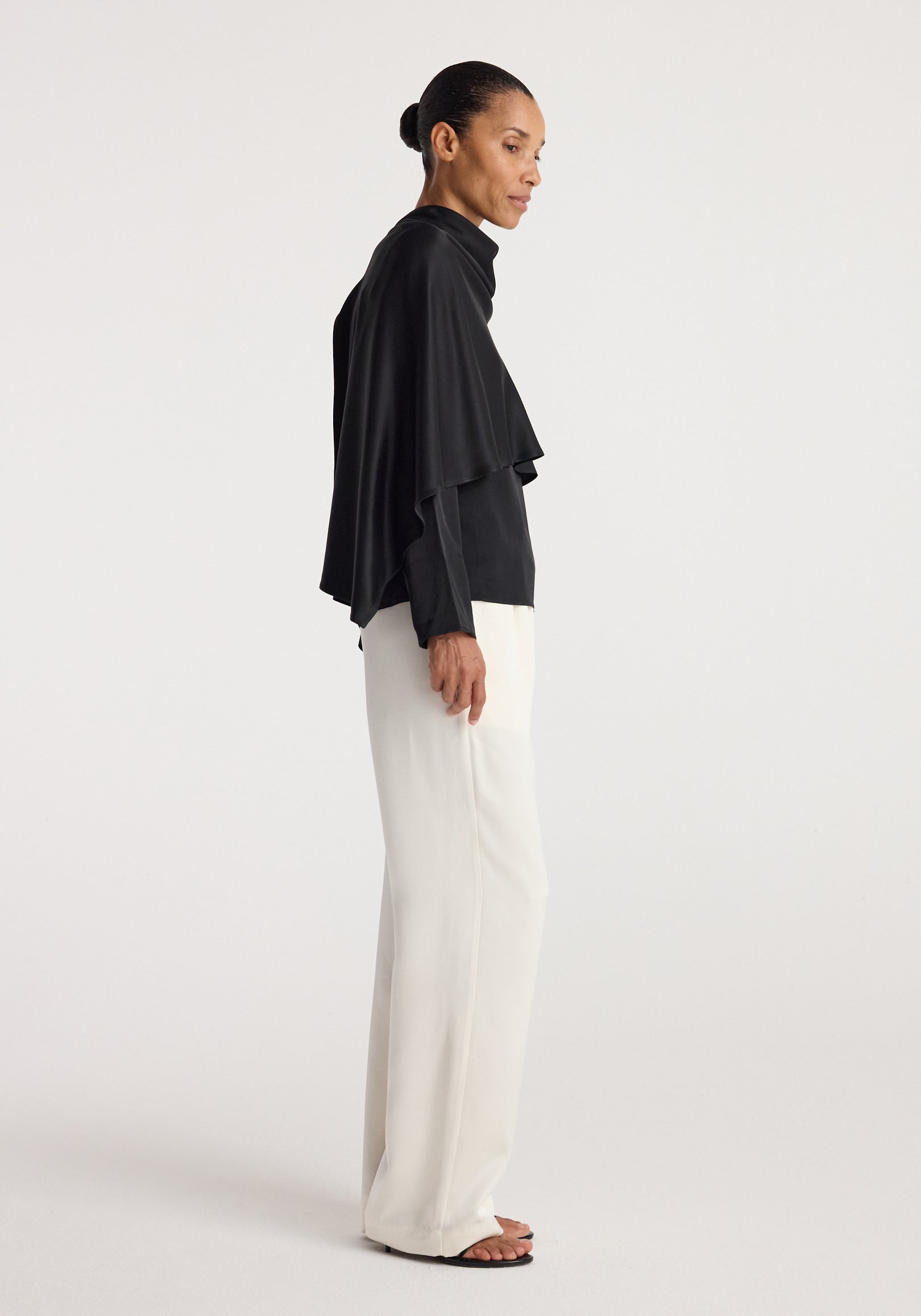Woman wearing a black Satin cape top | black paired with white wide-leg pants and black sandals, standing sideways against a plain background.