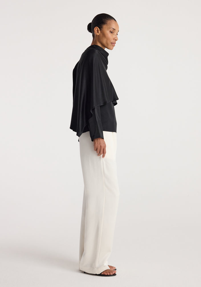 Woman wearing a black Satin cape top | black with a draped silhouette paired with loose white trousers and black open-toe sandals against a plain background.