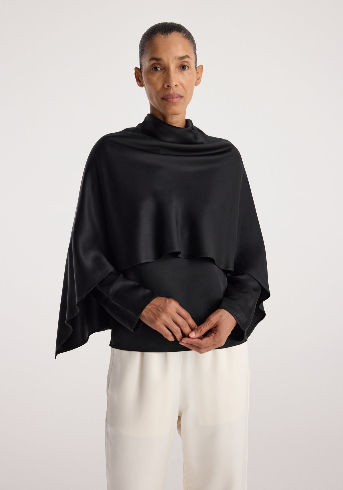 Woman wearing a black Satin cape top | black with a smooth, flowing texture, paired with white pants against a plain light background.