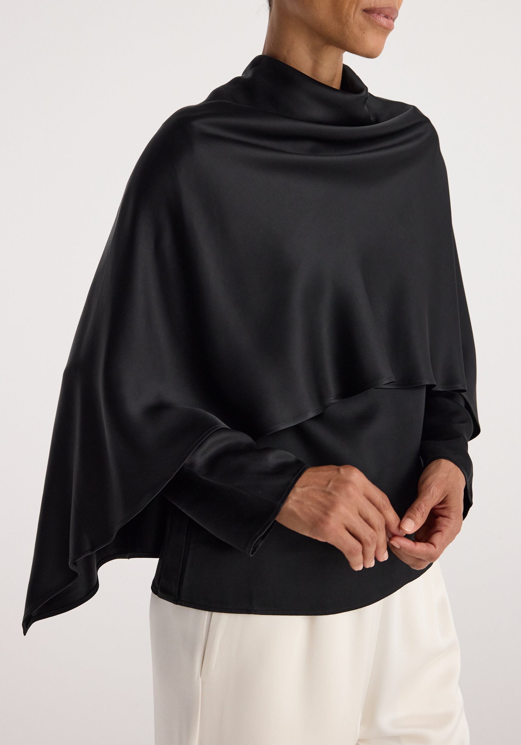 Woman wearing a black Satin cape top | black with a smooth, shiny texture, styled over a black long-sleeve shirt and white pants, shown from the waist up.