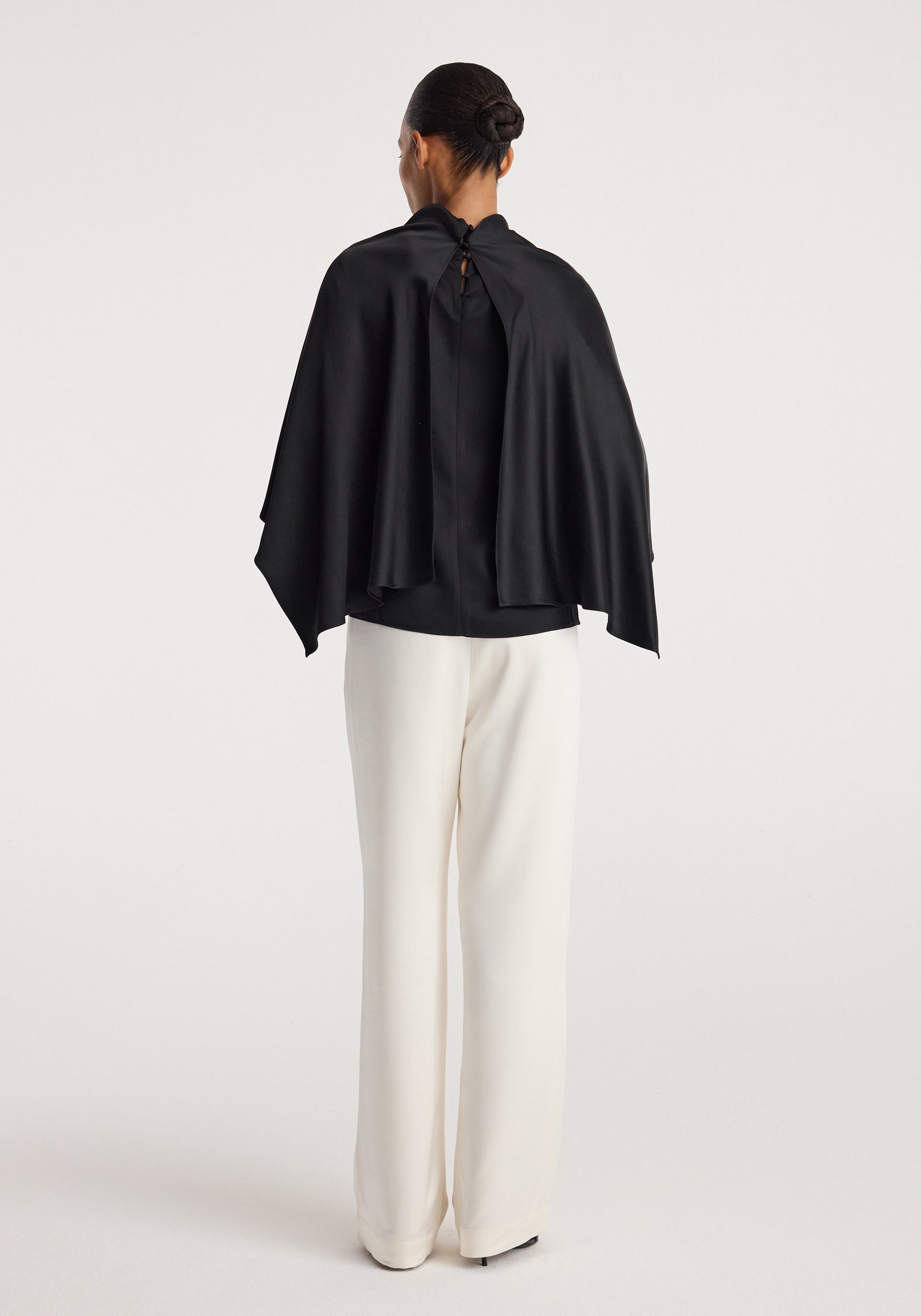 Woman wearing a black Satin cape top | black with a draped back detail and loose fit, paired with white wide-leg pants, standing against a plain light background.