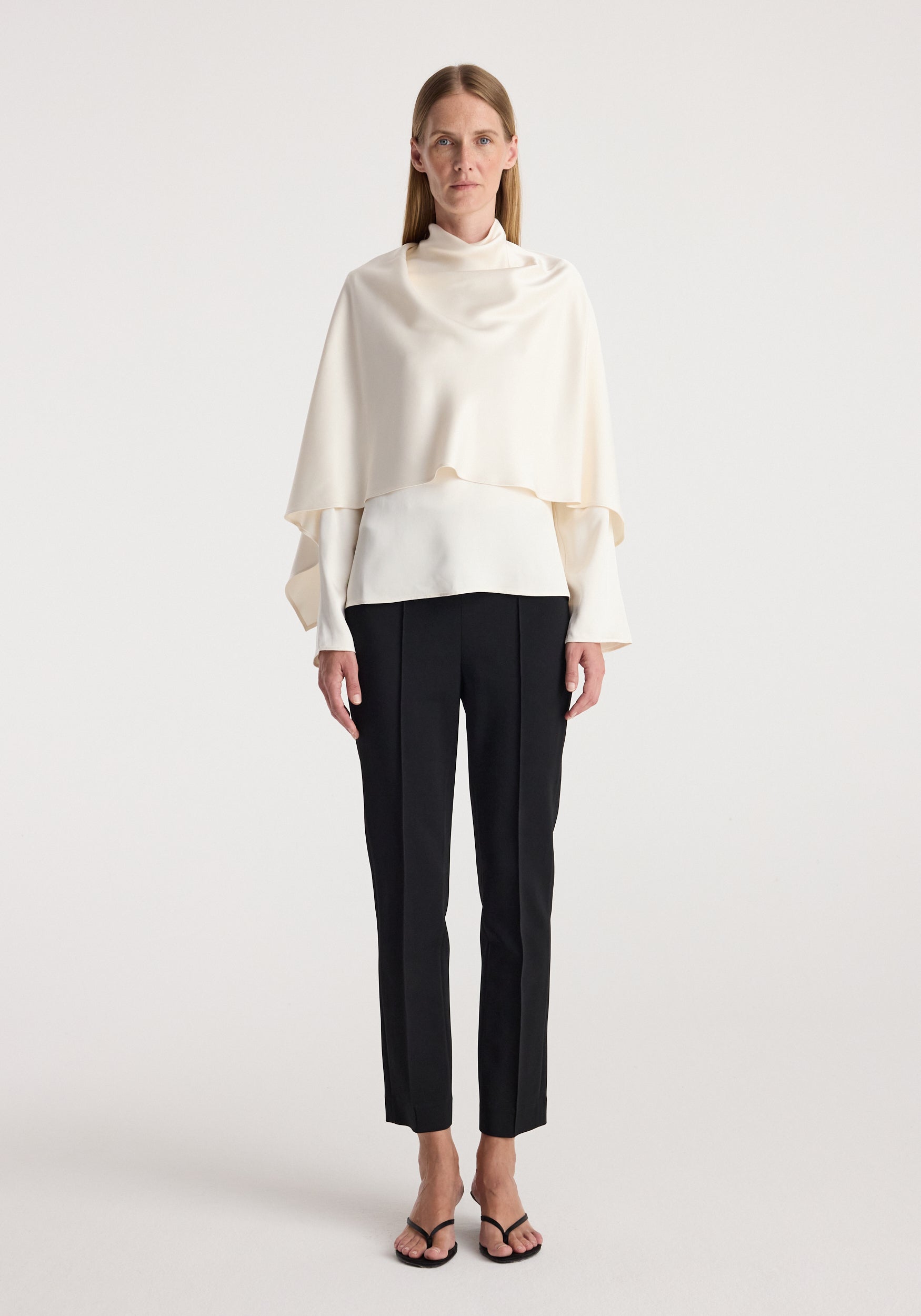 Woman wearing a cream satin cape top with long sleeves paired with black tailored pants and open-toe heels standing against a plain white background.